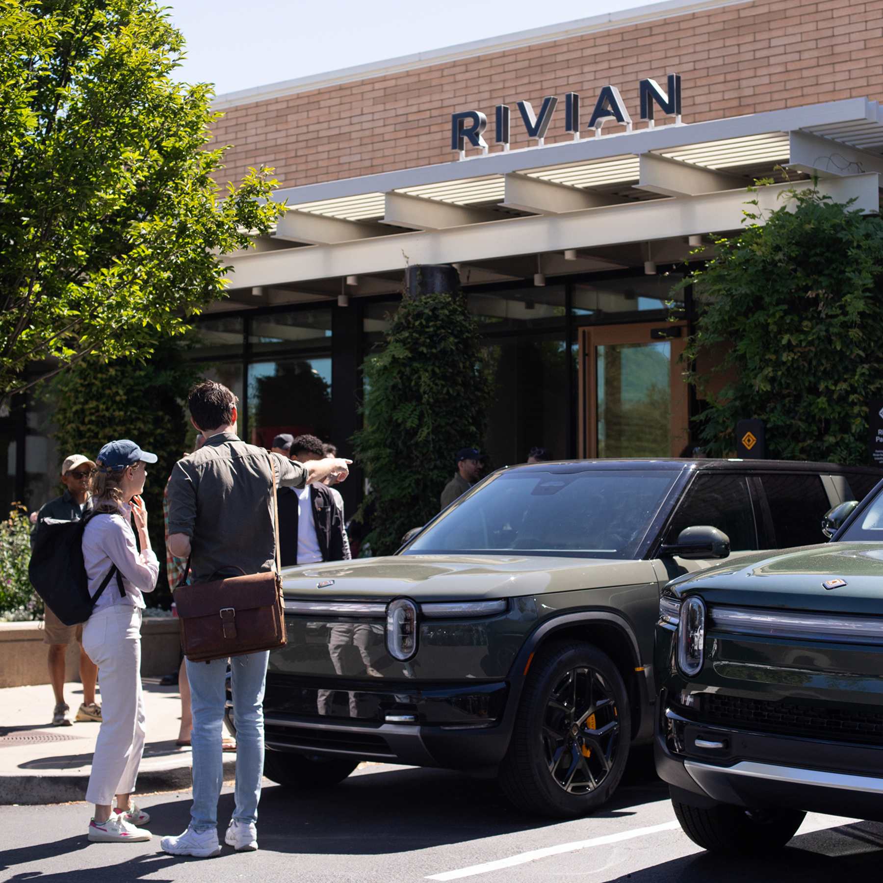 Two people seen from behind, looking at a white Rivian R2 SUV parked during a golden hour sunset.