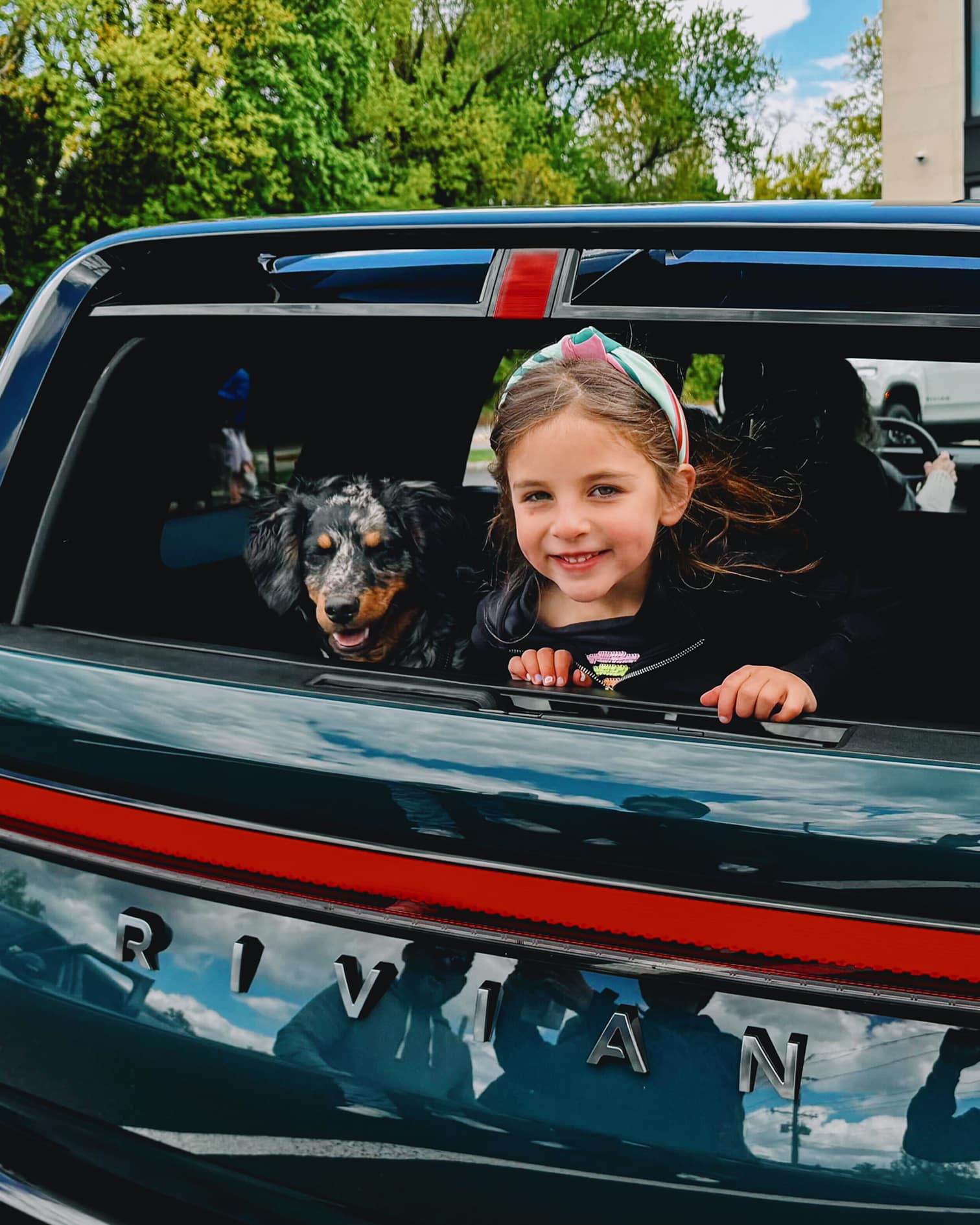 A smiling girl and her dog lean out of the open rear window of an R2 electric SUV at a Rivian R2 Block Party.