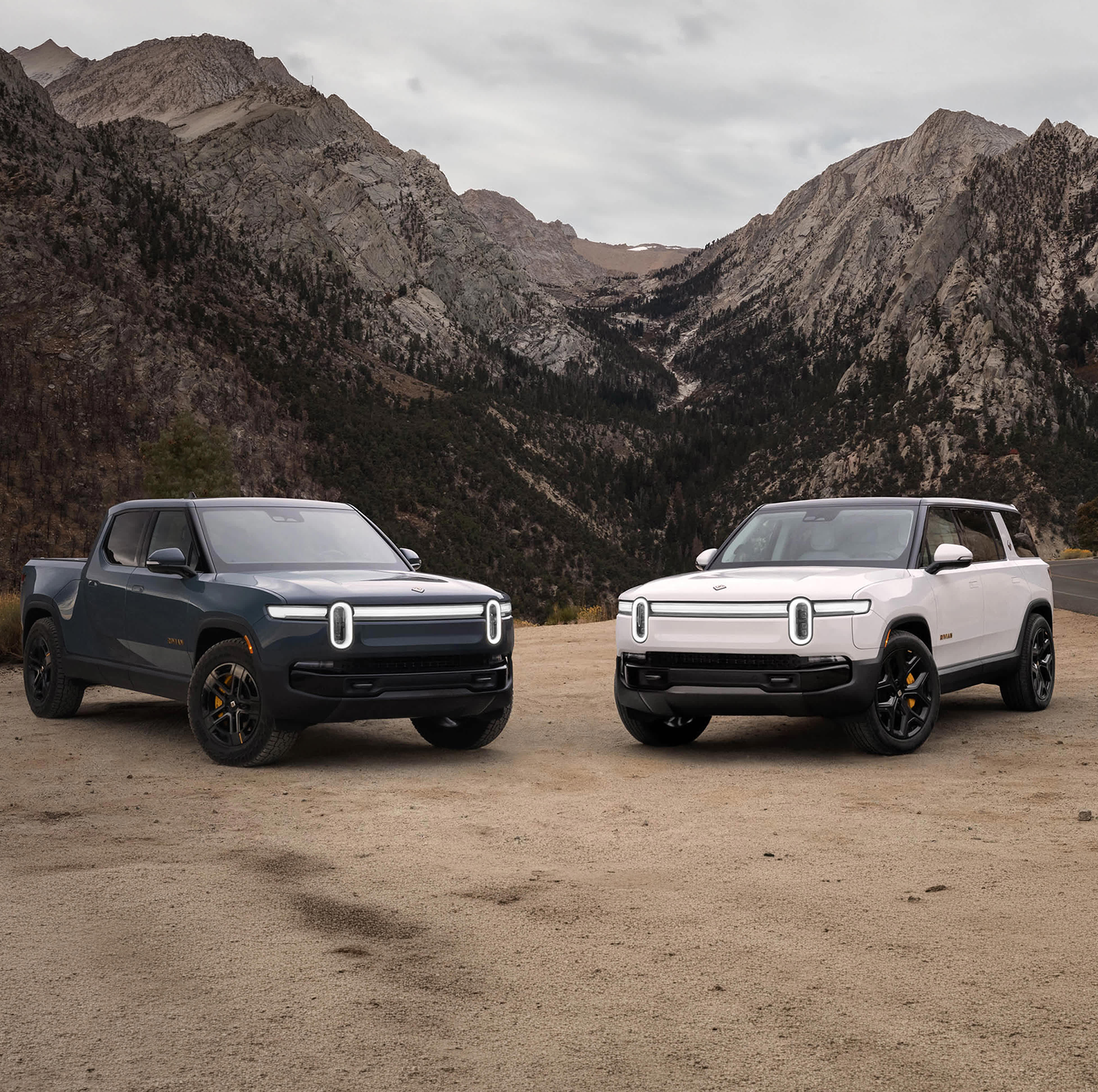 A storm blue rRivian R1t. electric truck and a white rivian R1S electric SUV parked on lookout along a winding highway on the side of a picture-esque mountain.