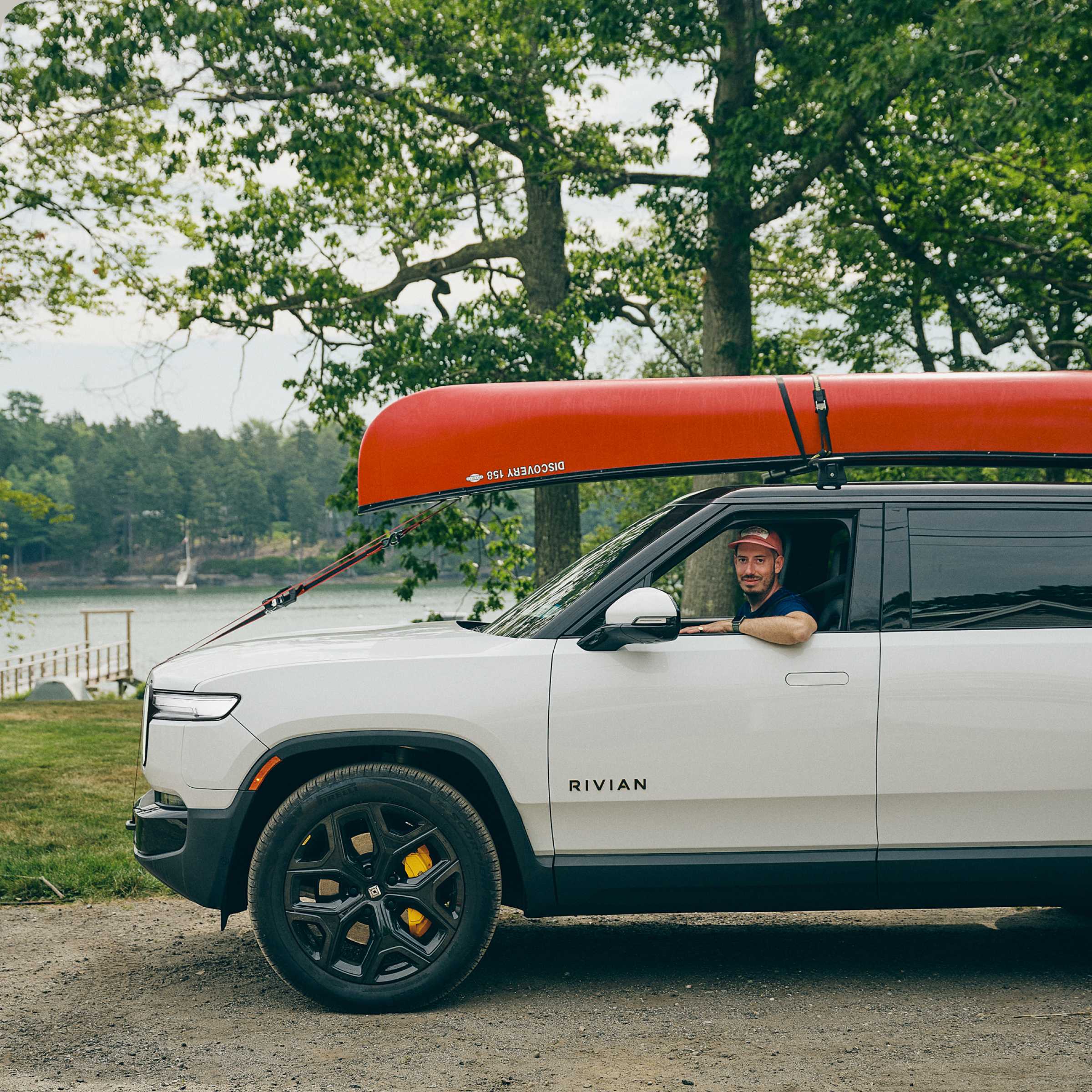 Side view of drive side of a white Rivian electric vehicle parked beside a tree lined lake. An orange canoe is strapped to the roof racks, and the driver is smiling while looking out the window