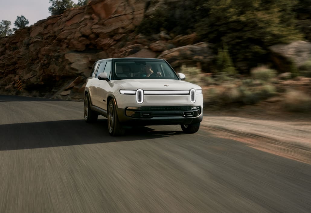 The front of a white Rivian Quad motor electric vehicle, swiftly driving down a open road with red-rock cliffs in the background.