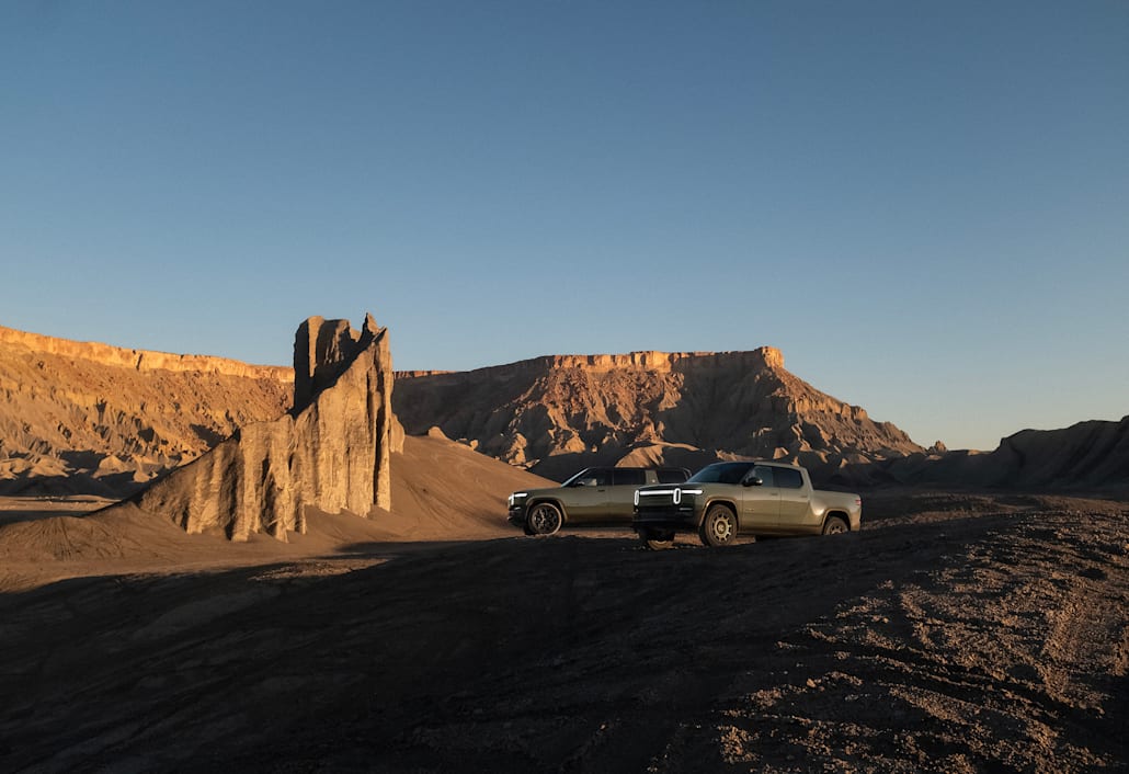 Two Rivian electric vehicles parked in a canyon with red-rock cliffs extensively filling the background. One car is a Rivian R1S electric SUV an the other is Rivian R1T electric SUV. Both are in the limited edition Launch Green color.