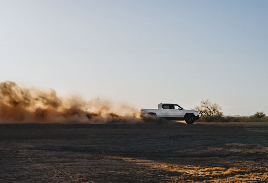 A white Rivian R1T electric truck driving swiftly through a dirt valley as the sun sets. A plume of dust kicks out of the back.