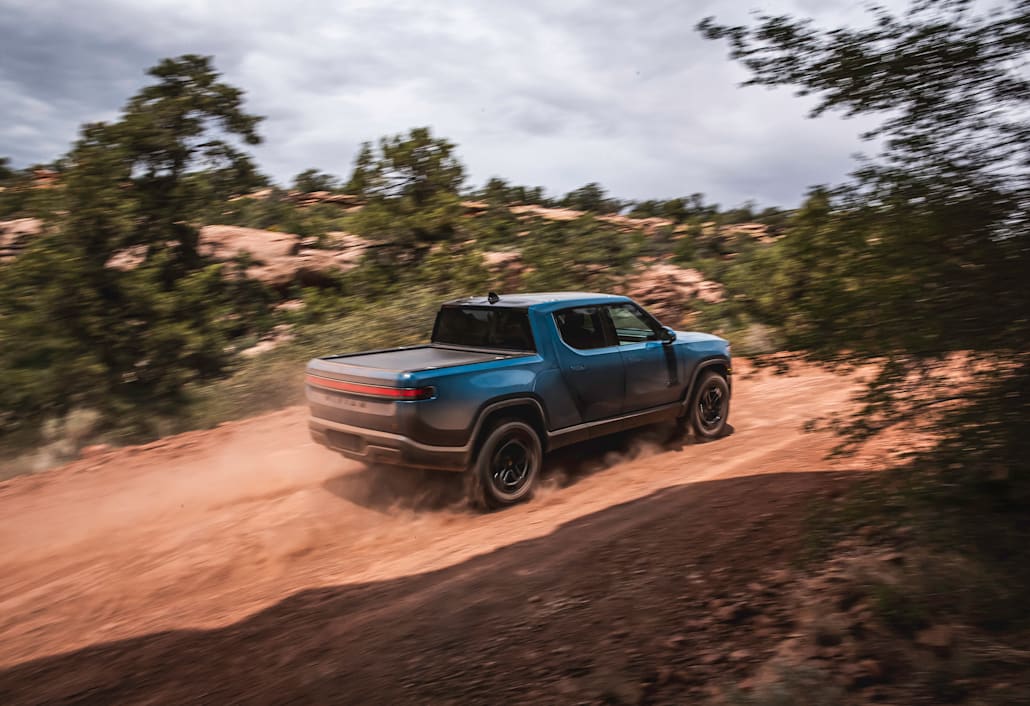 A backside view of a blue Rivian R1T electric truck driving up a dusty dirt road.