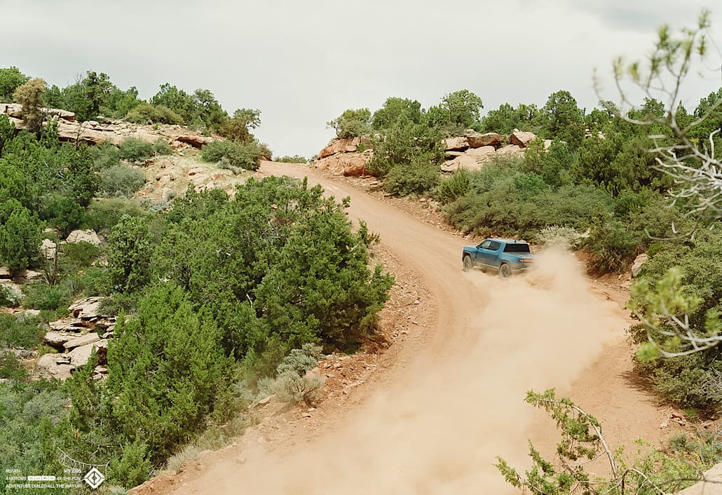 A Rivian R1T electric truck drifting a left turn as it drives up a dusty dirt road.