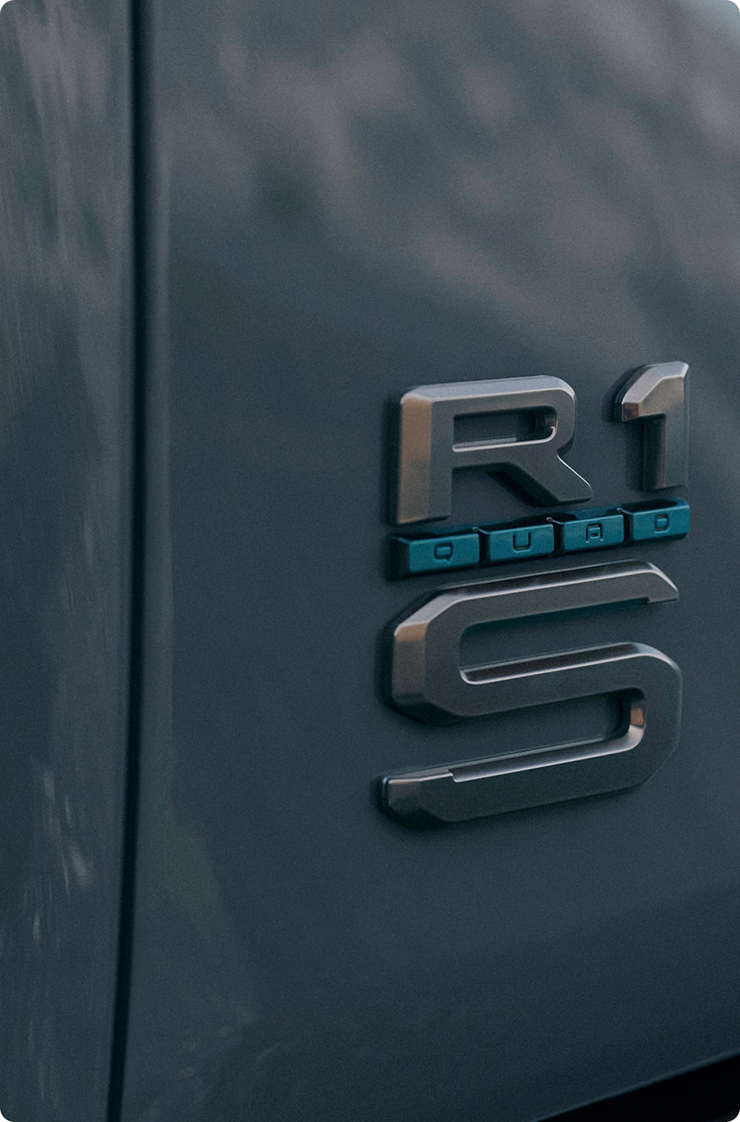 The rear truck badging of a Rivian Quad motor electric vehicle. The badge shows the R1 logo, but featuring a "QUAD" badge trimmed with Laguna Blue accents, exclusive to Quad.
