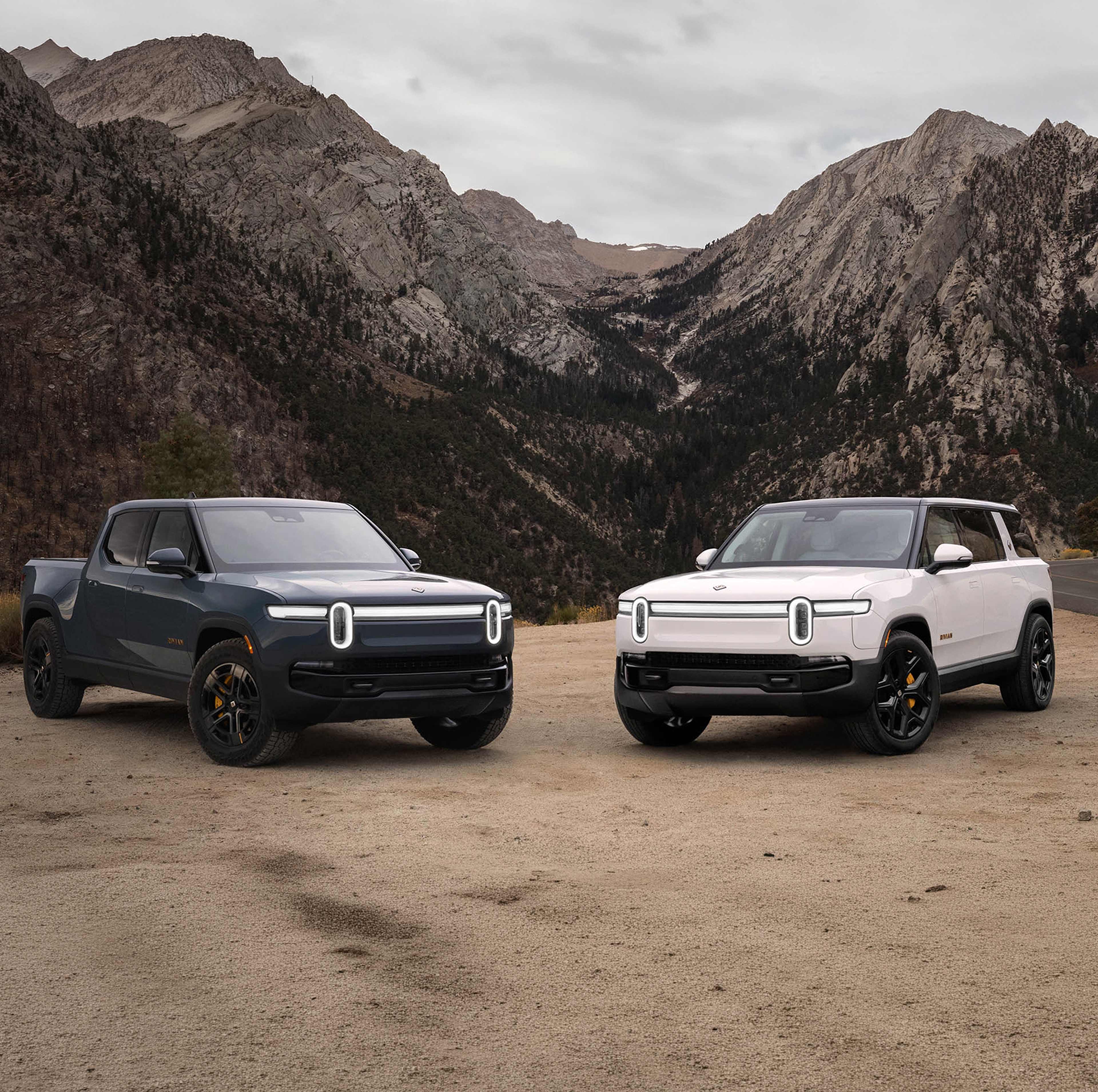 A storm blue rRivian R1t. electric truck and a white rivian R1S electric SUV parked on lookout along a winding highway on the side of a picture-esque mountain.