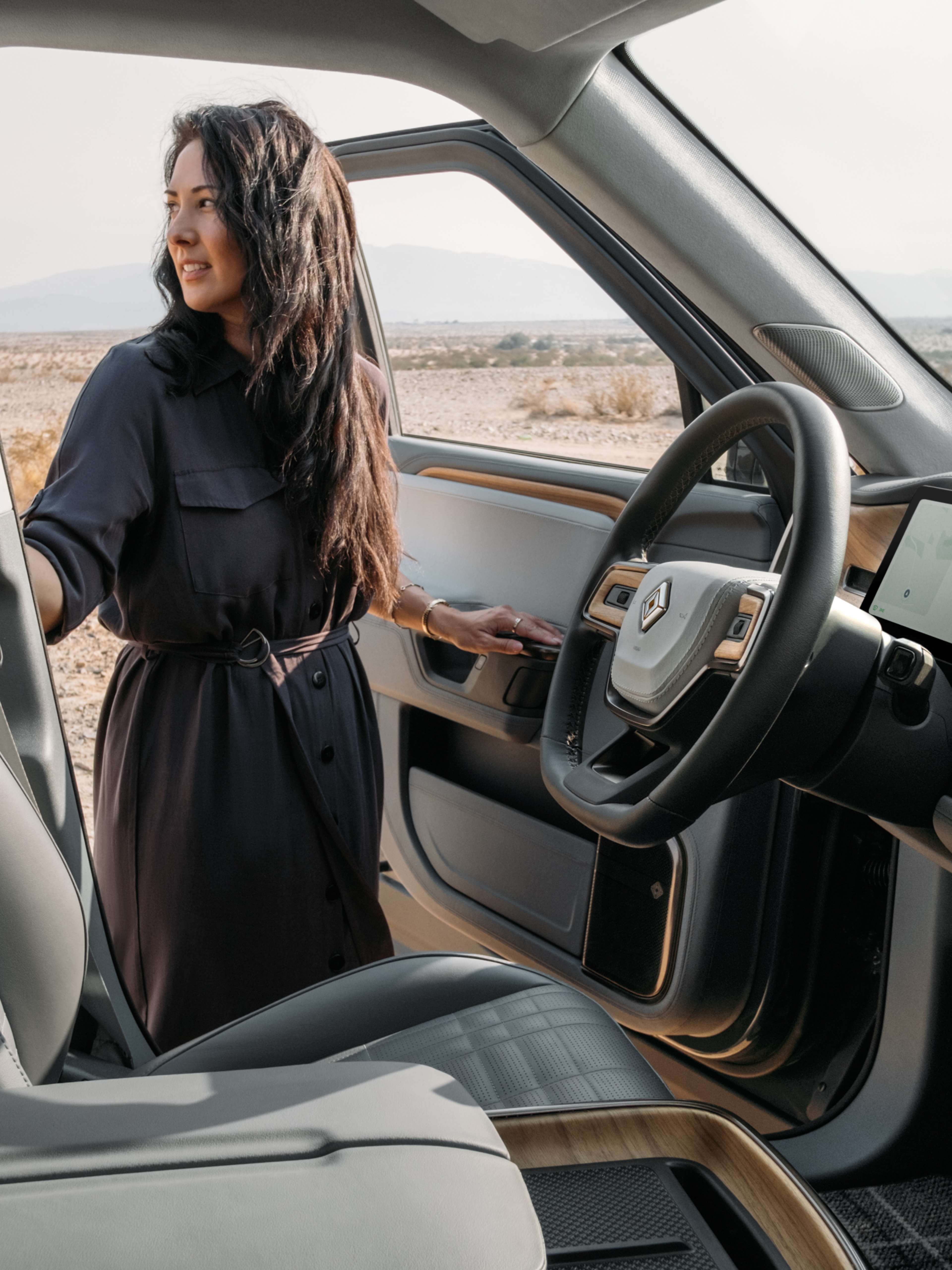 A woman in a dark dress standing beside the open driver door of a Rivian EV car.