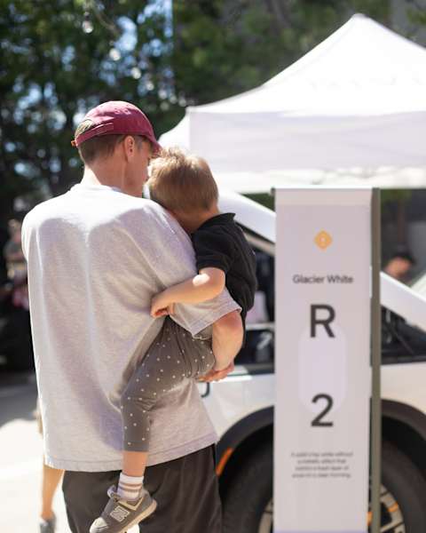 A father holding a toddler stands next to a "Glacier White R2" display banner at a Rivian R2 Block Party.
