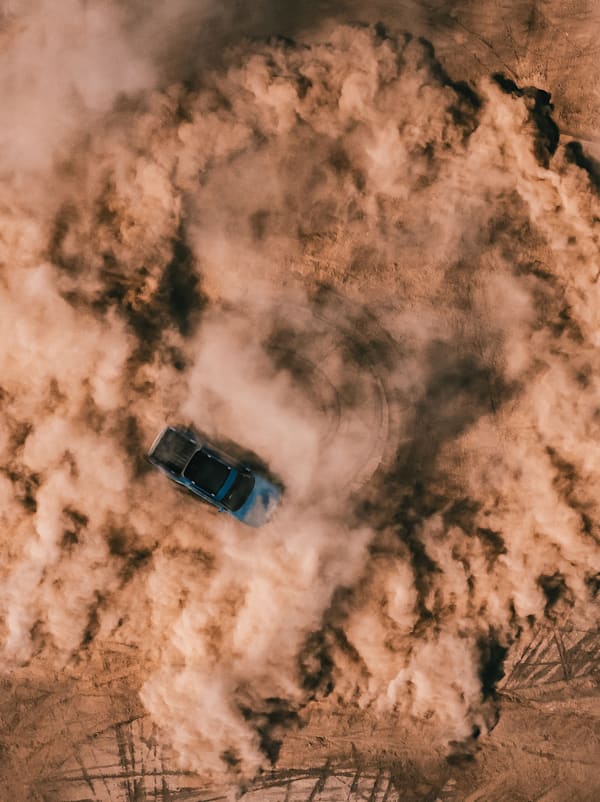 An aerial shot of a Rivian Quad motor electric vehicle utilizing 4 motor vectoring to perform a tight, controlled, circular spin on a dirt landscape. Dust fills the air and circular tire marks line the ground.