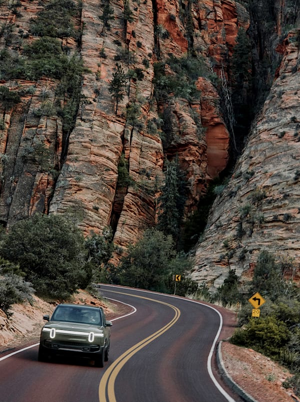 The front of a Rivian Quad motor electric vehicle driving down a winding road. A large rocky cliff is in the background.