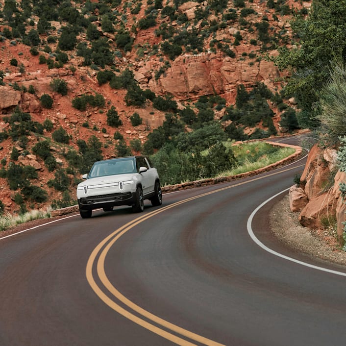 The front of a white Rivian R1S Quad motor electric SUV driving through down a narrow-winding highway with red rocks cliffs in the background.