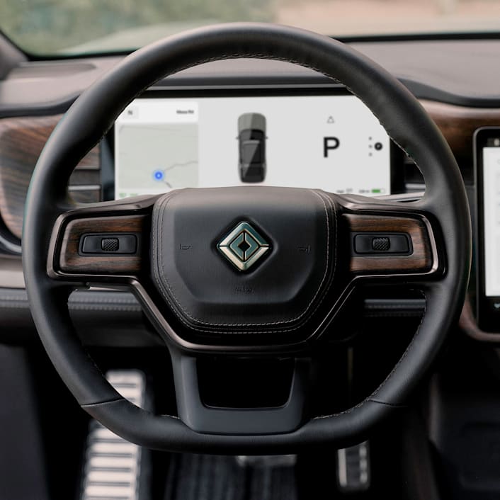 The steering wheel and odometer screen of a Rivian electric vehicle. The black leather steering wheel is trimmed with dark wood accents.