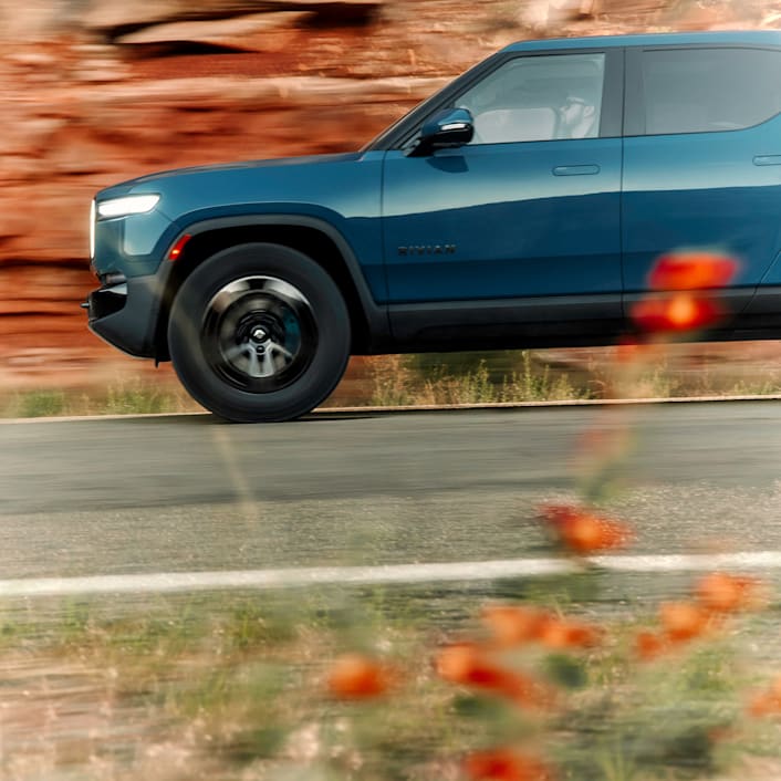 A cutoff view of the front driver side of a Rivian electric car. The car features Laguna Blue paint color. Red rock cliffs are in the background.