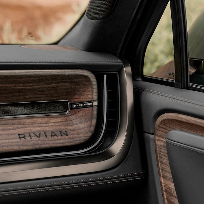 An interior view of the passenger glove box of a Rivian electric vehicle. The passenger glove compartment and dashboard are lined with black leather and features dark wood accents.