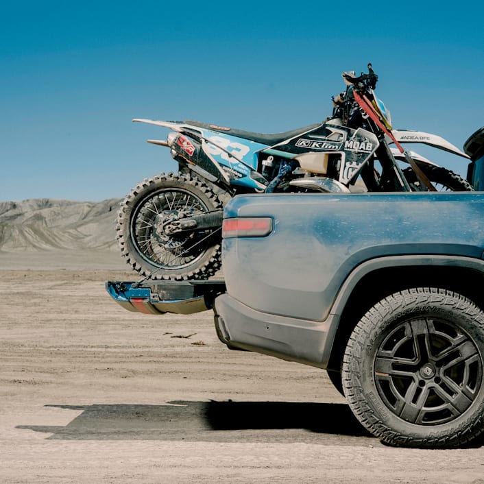 A cut-off image of the back of a Rivian R1T electric truck, covered in dust. The trunk is carrying a dirt bike.