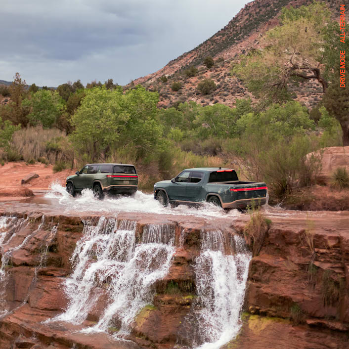 Two Rivian electric vehicles driving through a heavy stream of water atop a red-rock cliff. One car is a Rivian R1S electric SUV with Forest Green paint, and the other is Rivian R1T electric SUV with Storm Blue paint.