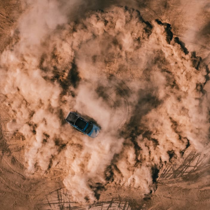 An aerial shot of a Rivian Quad motor electric vehicle utilizing 4 motor vectoring to perform a tight, controlled, circular spin on a dirt landscape. Dust fills the air and circular tire marks line the ground.