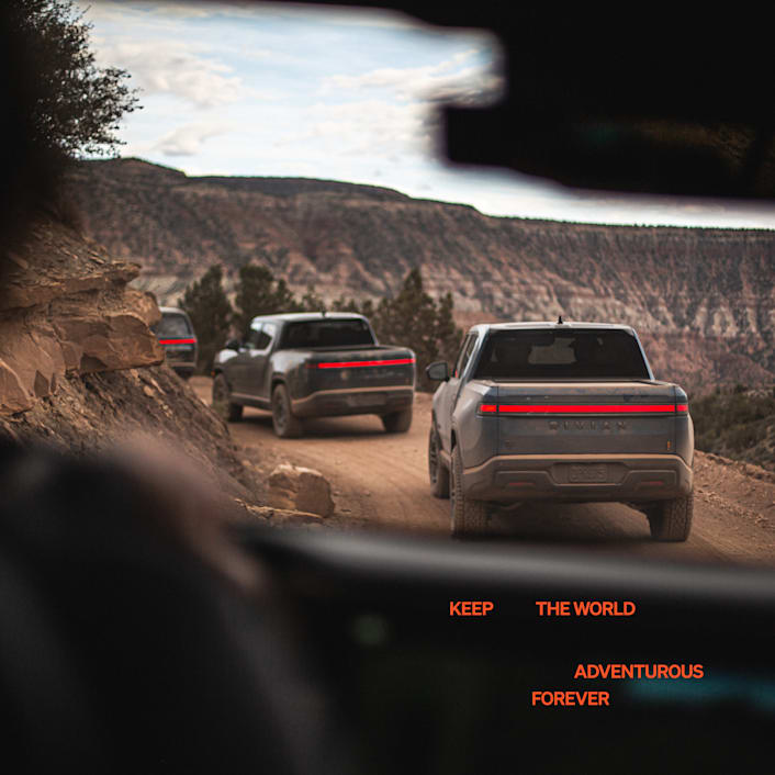 A line of three Rivian R1T electric trucks driving around a winding dirt road. The picture is taken from the inside of another Rivian vehicle that is trailing these three vehicles. 