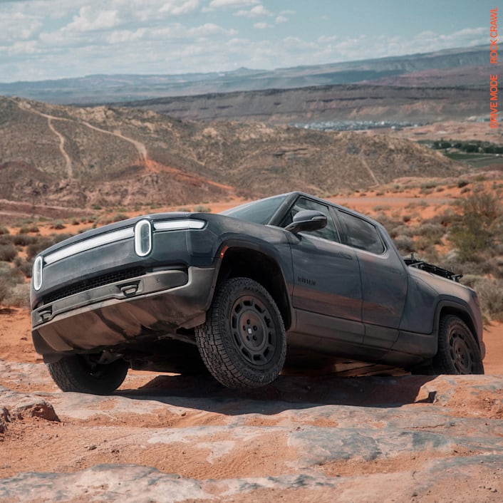 A Rivian R1T electric truck crawling over rugged terrain. The front driver wheel is off the ground. The car features 20” Dune Satin Graphite All-Terrain wheels. 