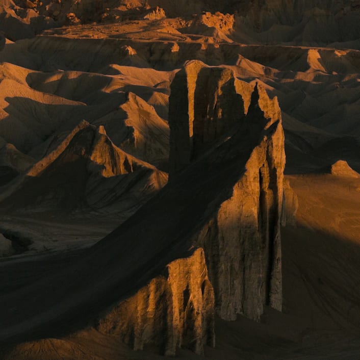 A massive slanted red rock cliff, with numerous other smaller hills in the background. The cliffs cast shadows in the sundown light.