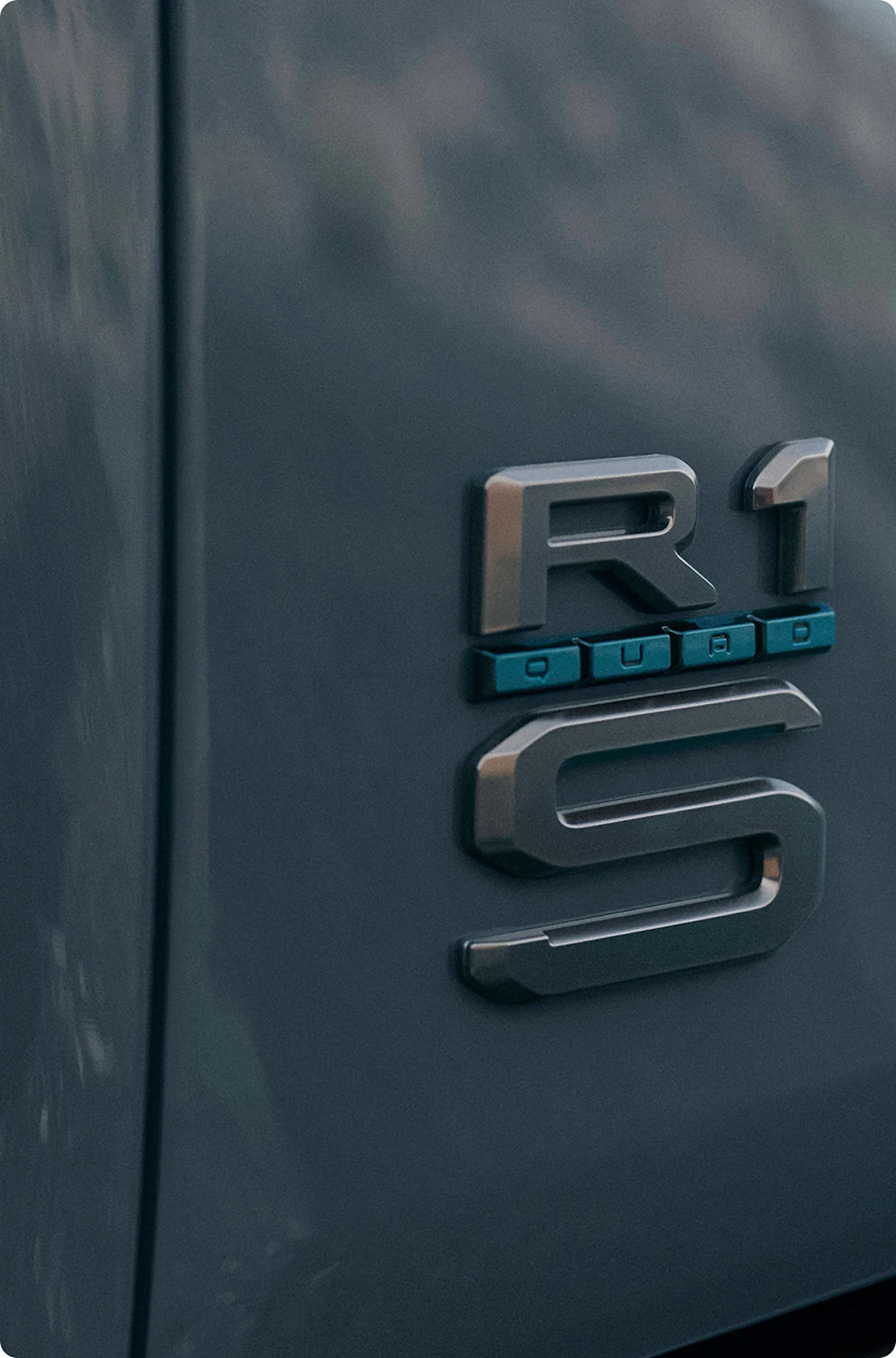 The rear truck badging of a Rivian Quad motor electric vehicle. The badge shows the R1 logo, but featuring a "QUAD" badge trimmed with Laguna Blue accents, exclusive to Quad.