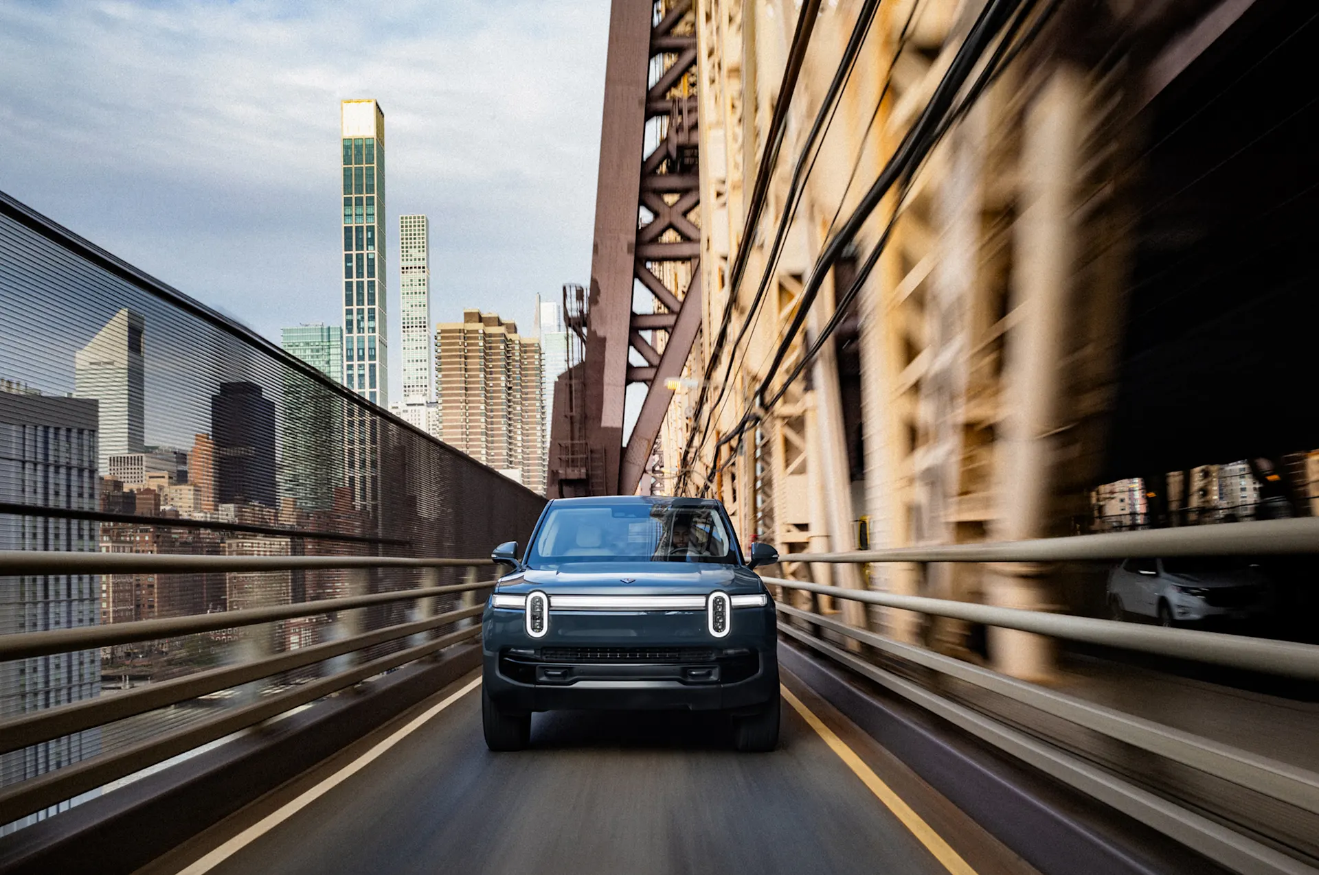 The front of a Rivian electric car driving across a New York City bridge. The support structure of the bridge and the high rise buildings fill the background.