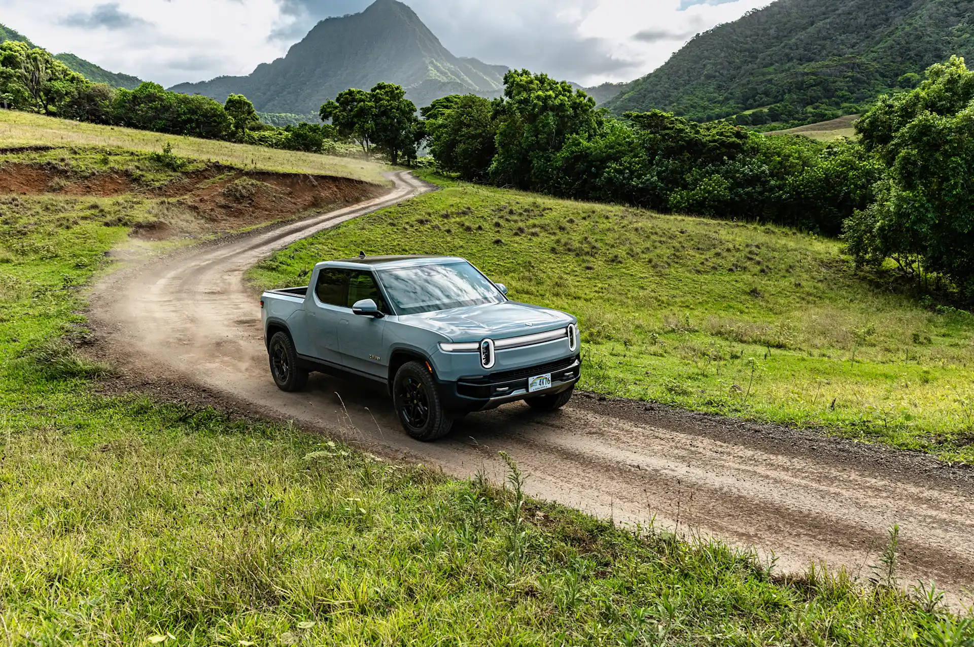A Rivian EV truck driving down a rural dirt road. Green foliage and green covered mountains are in the background.