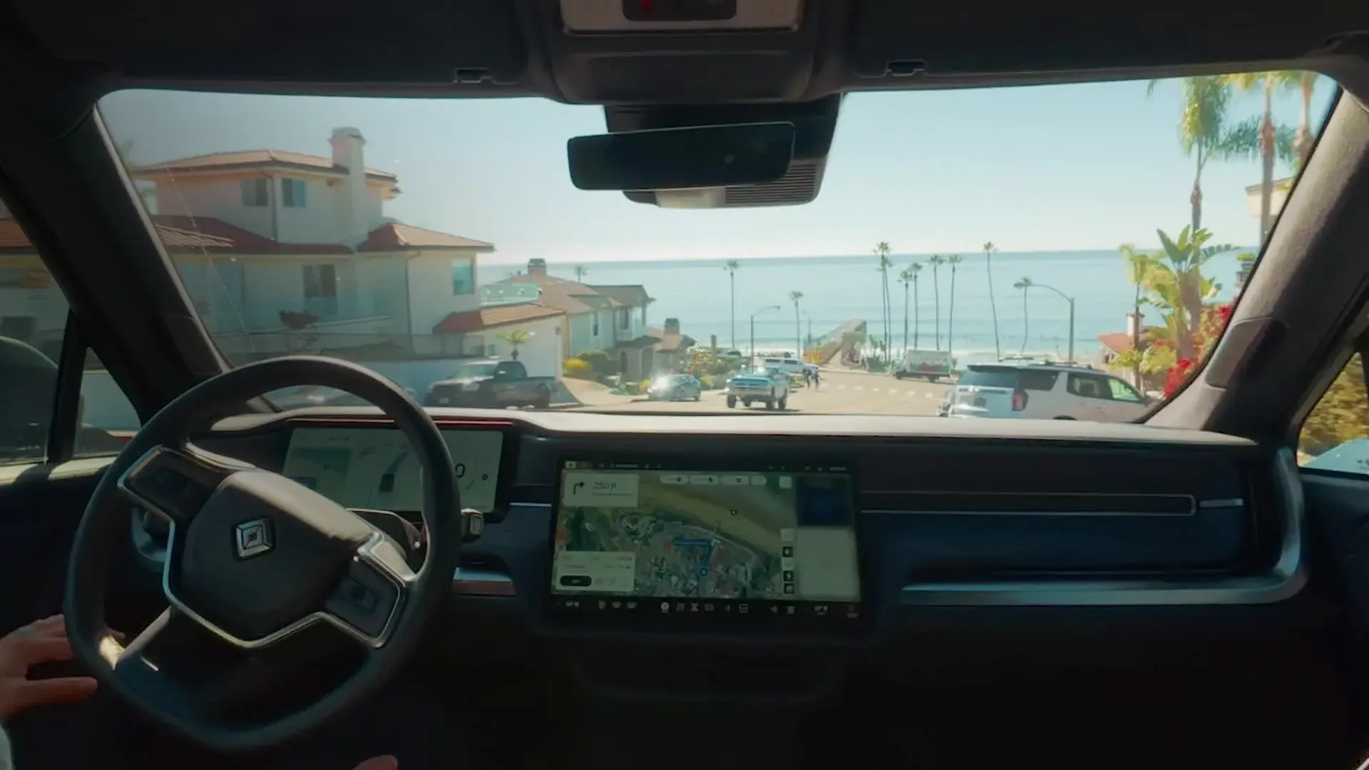Driver's view inside a Rivian EV, looking out at a sunny, palm-lined street sloping toward the ocean. The minimalist interior features a digital dashboard and a large central touchscreen displaying a navigation map.