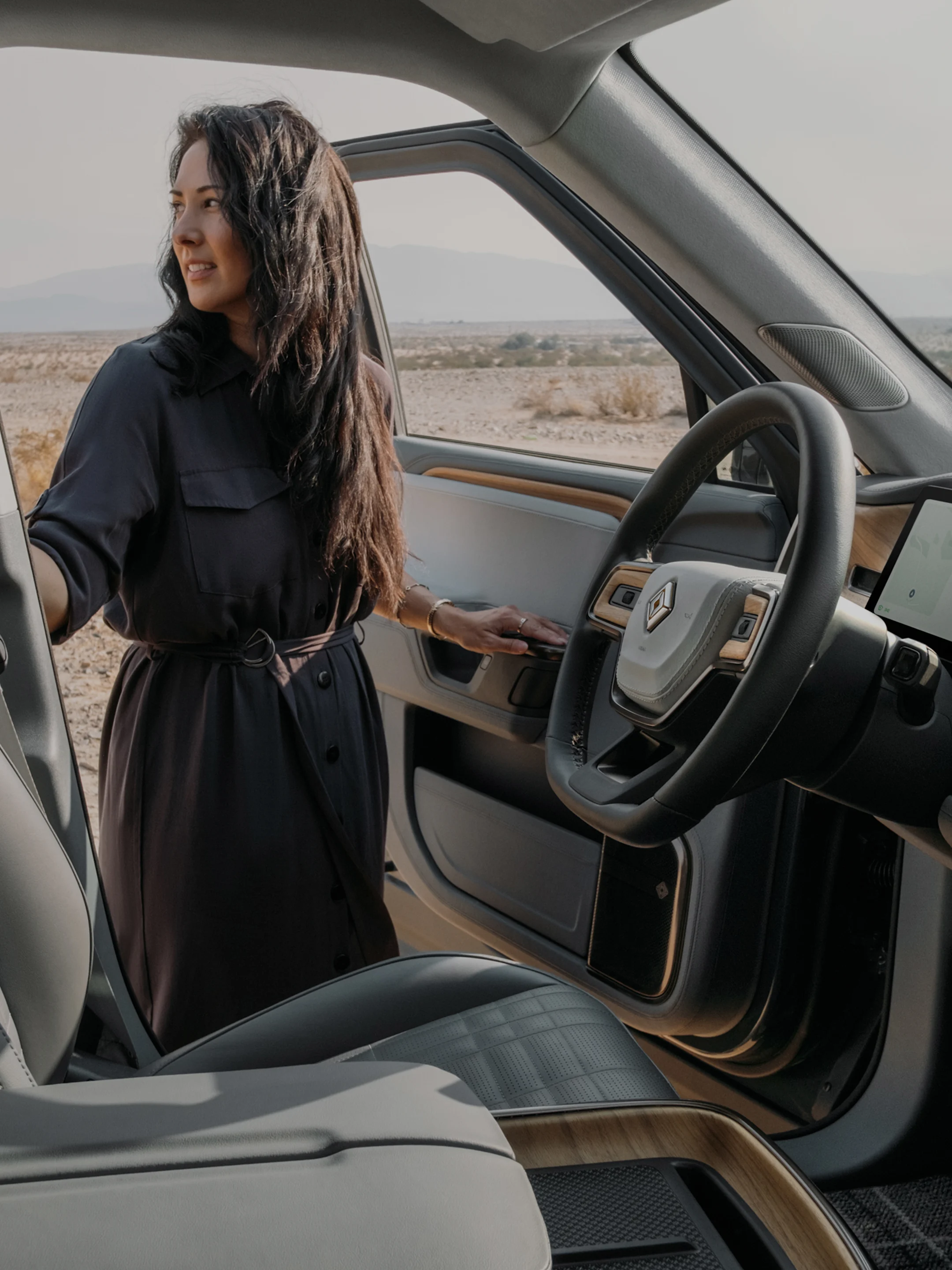 A woman in a dark dress standing beside the open driver door of a Rivian EV car.