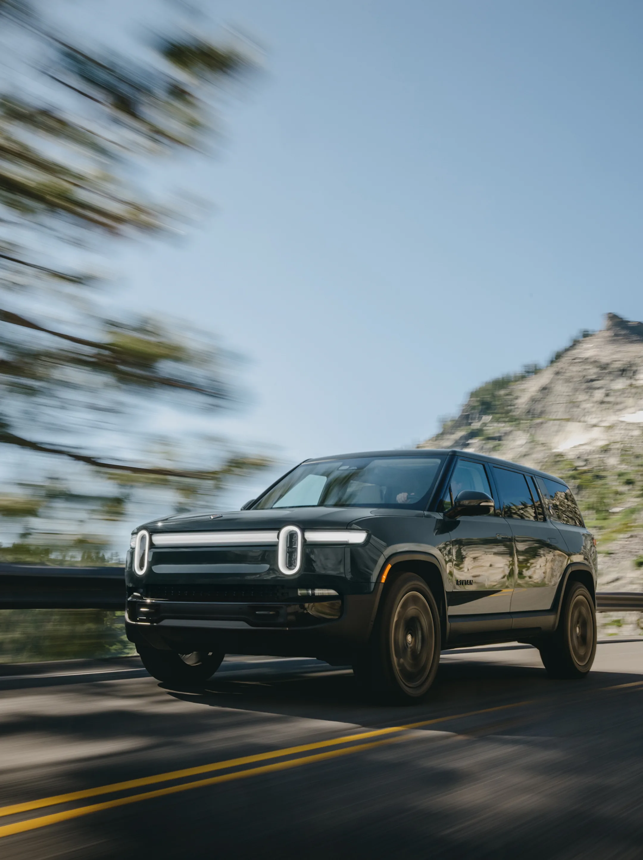 A dark colored Rivian Quad motor electric vehicle. This electric SUV has motion blue as it drives down a road with trees and mountain in the background.