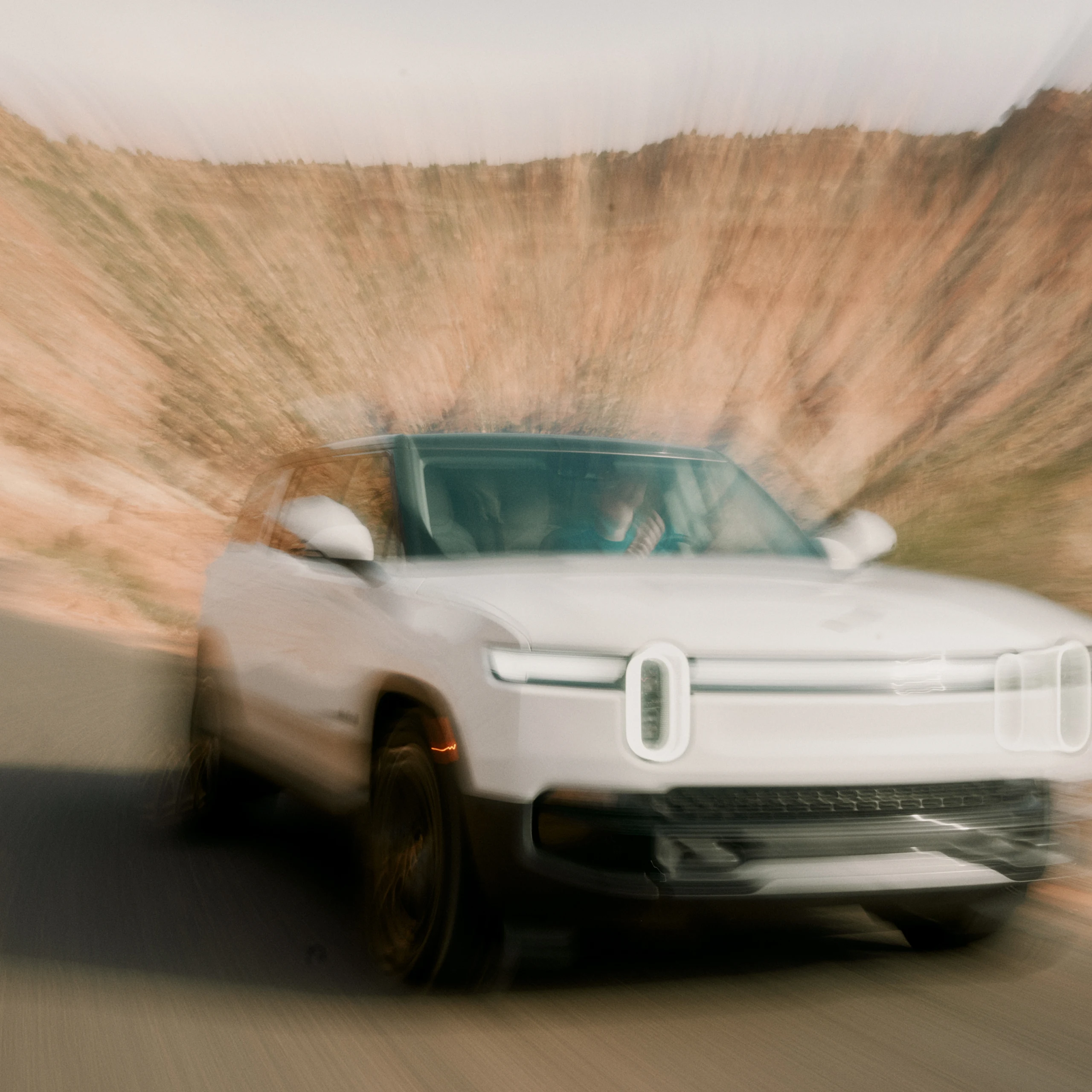 The front of a Rivian Quad motor electric vehicle driving down a winding road. A large rocky cliff is in the background.