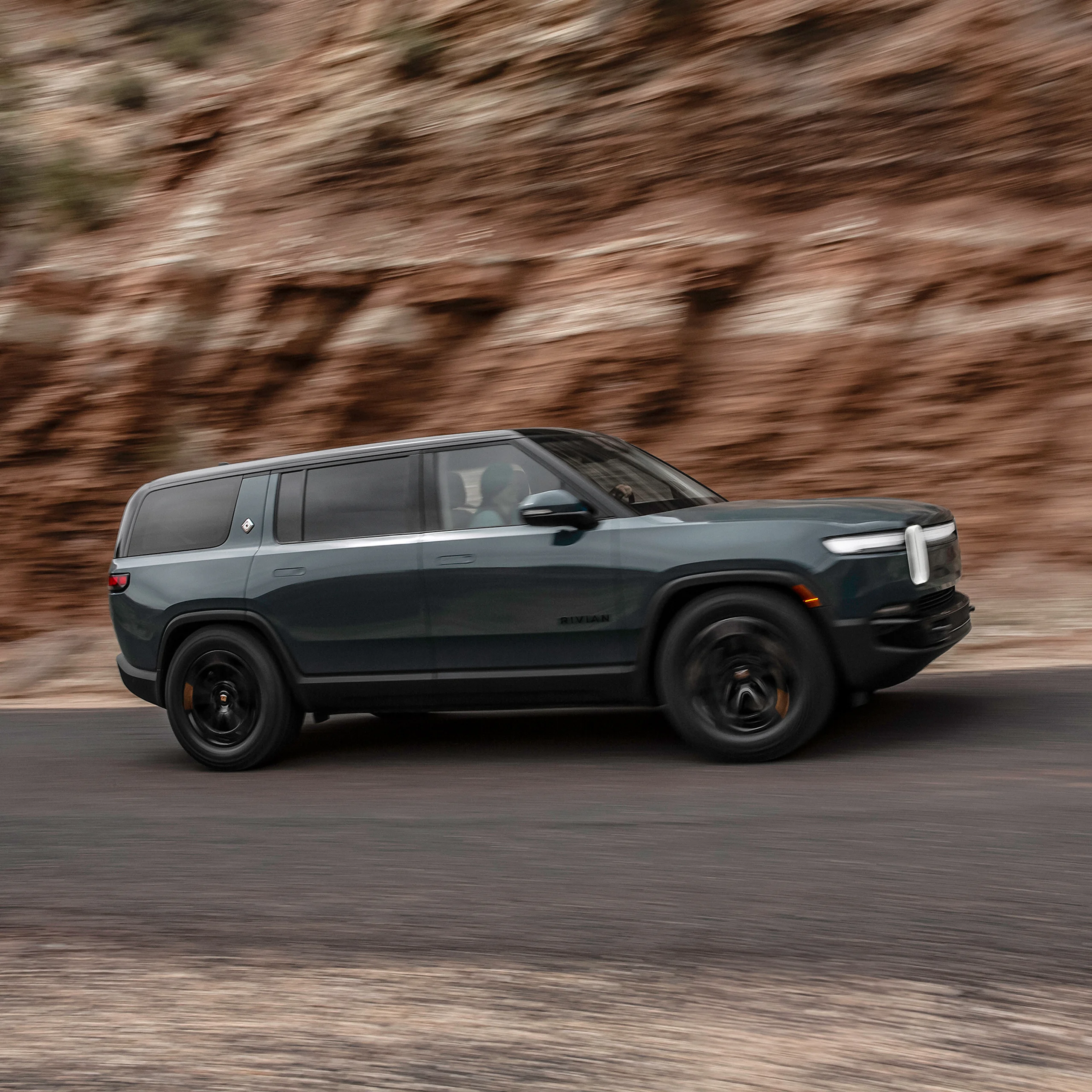 A dark grey colored Rivian R1S electric SUV driving swiftly up a road with red-rock cliffs close in the background. The car features dark wheels, dark badging, and Laguna Blue accents that are exclusive to Quad motor trims.