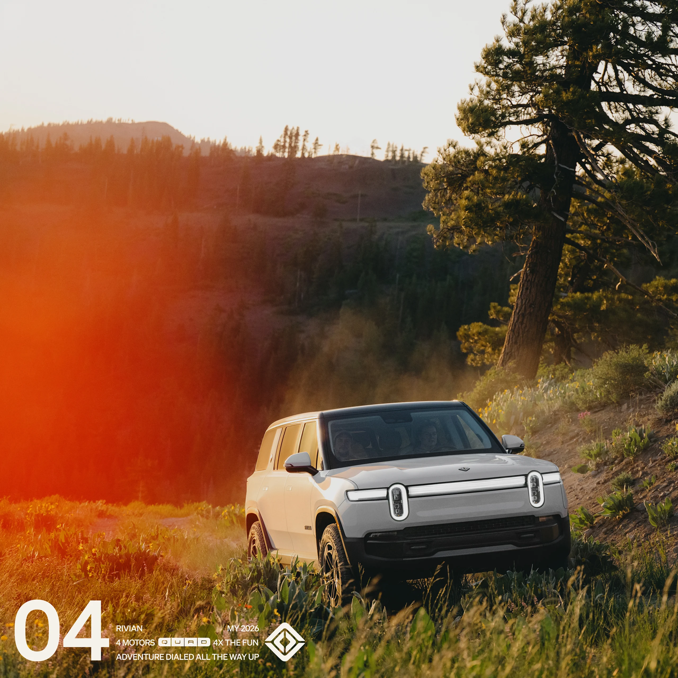 The front of a white Rivian R1S Quad motor electric SUV driving through grassy dirt road with tree-lined hills in the background.