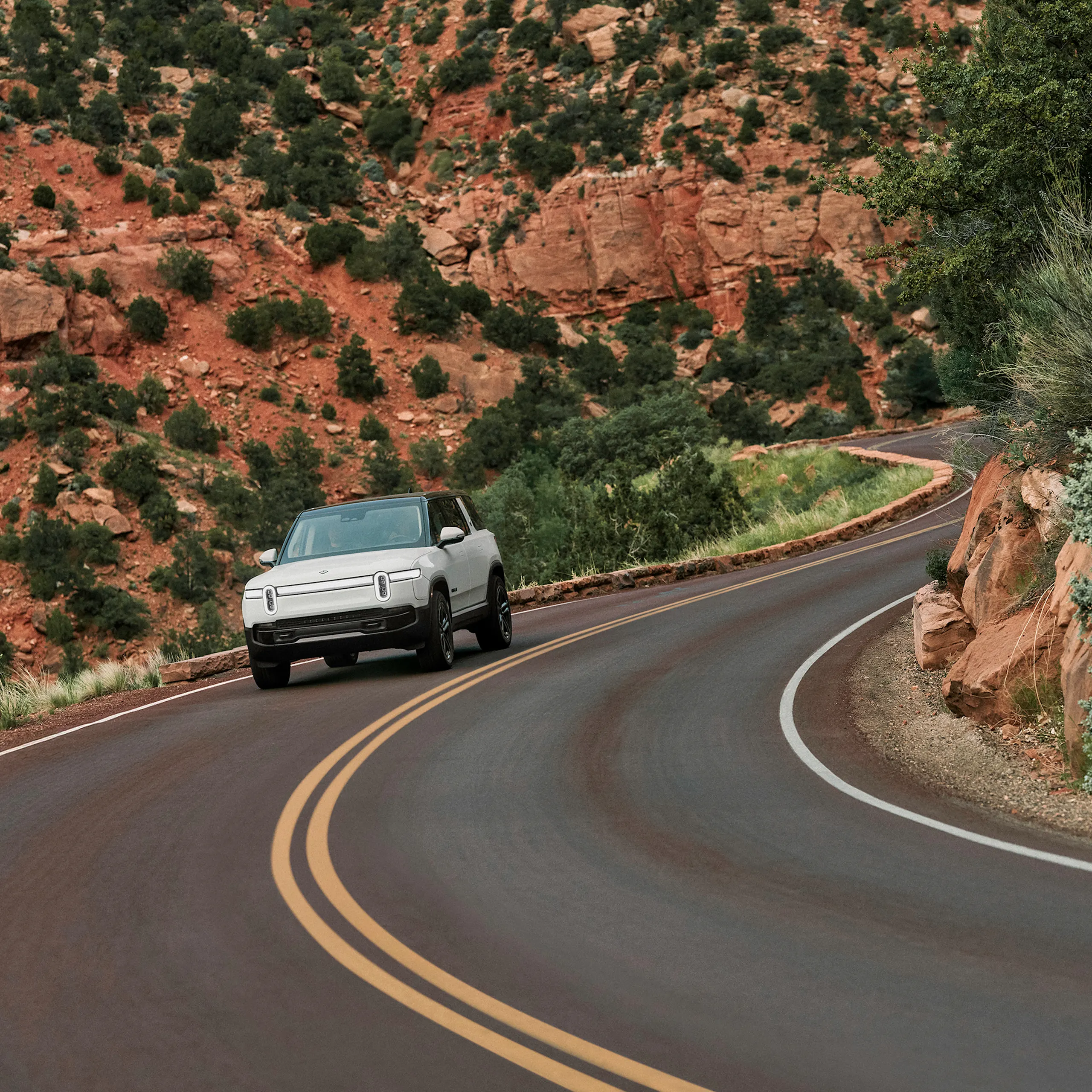 The front of a white Rivian R1S Quad motor electric SUV driving through down a narrow-winding highway with red rocks cliffs in the background.