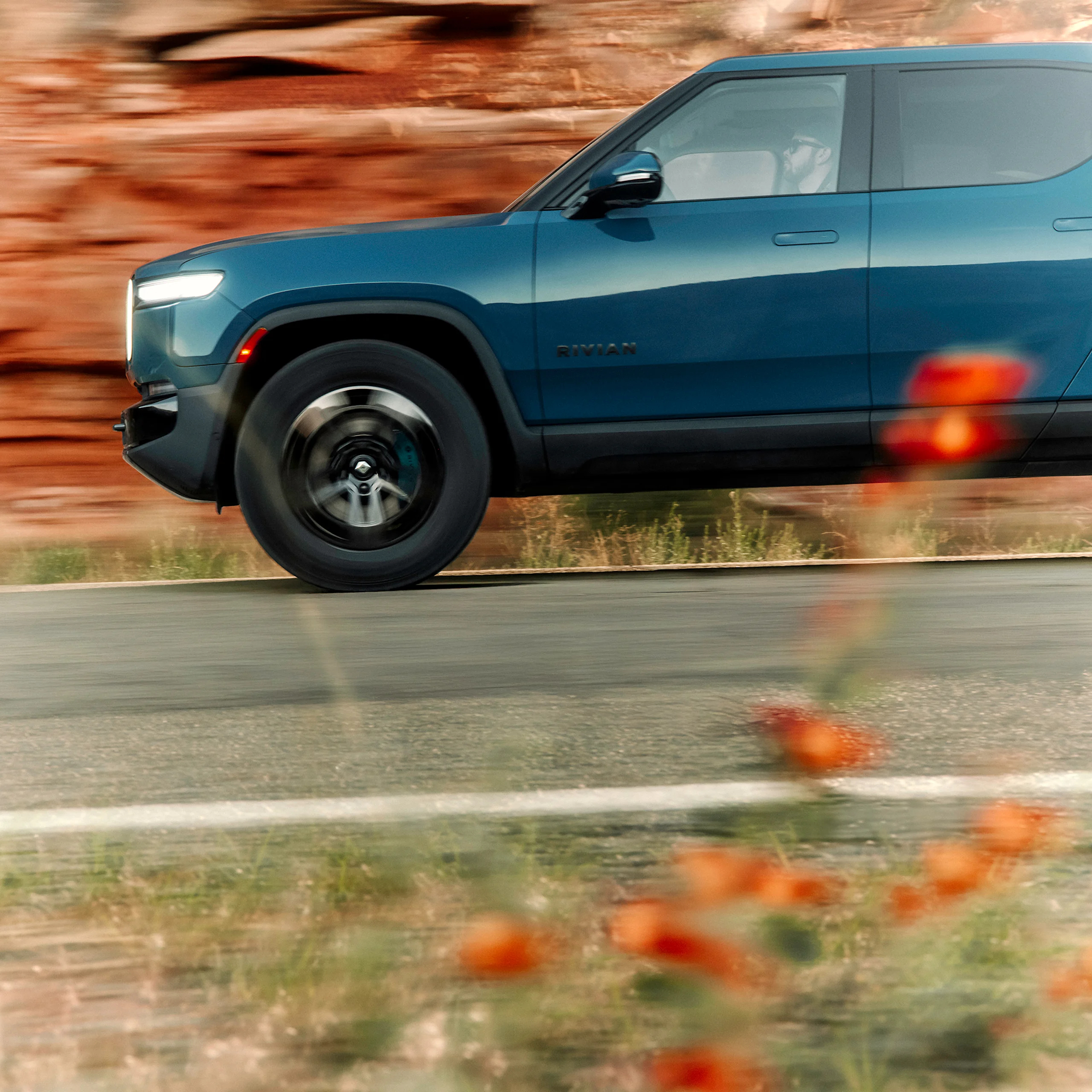 A cutoff view of the front driver side of a Rivian electric car. The car features Laguna Blue paint color. Red rock cliffs are in the background.