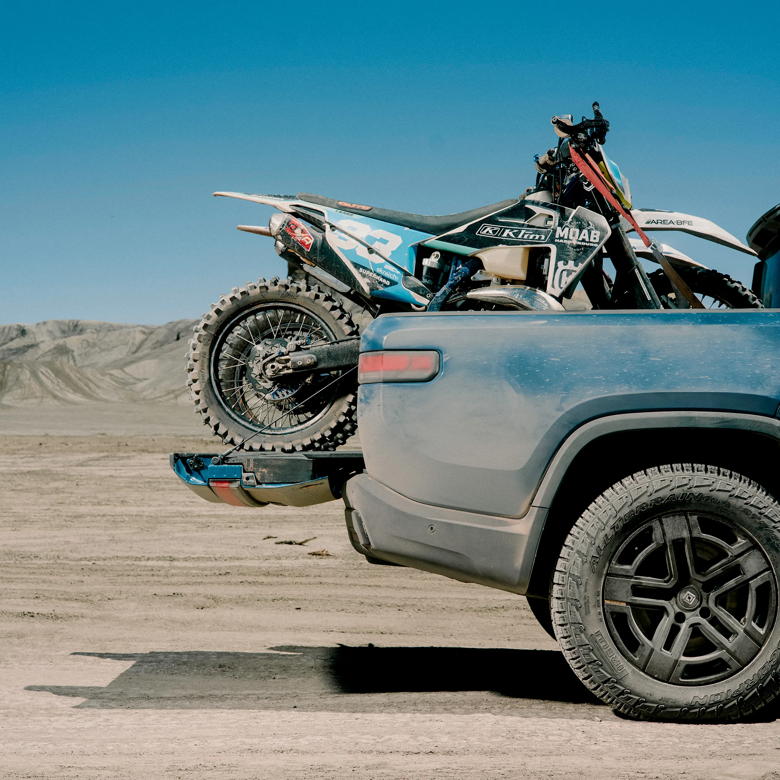 A cut-off image of the back of a Rivian R1T electric truck, covered in dust. The trunk is carrying a dirt bike.