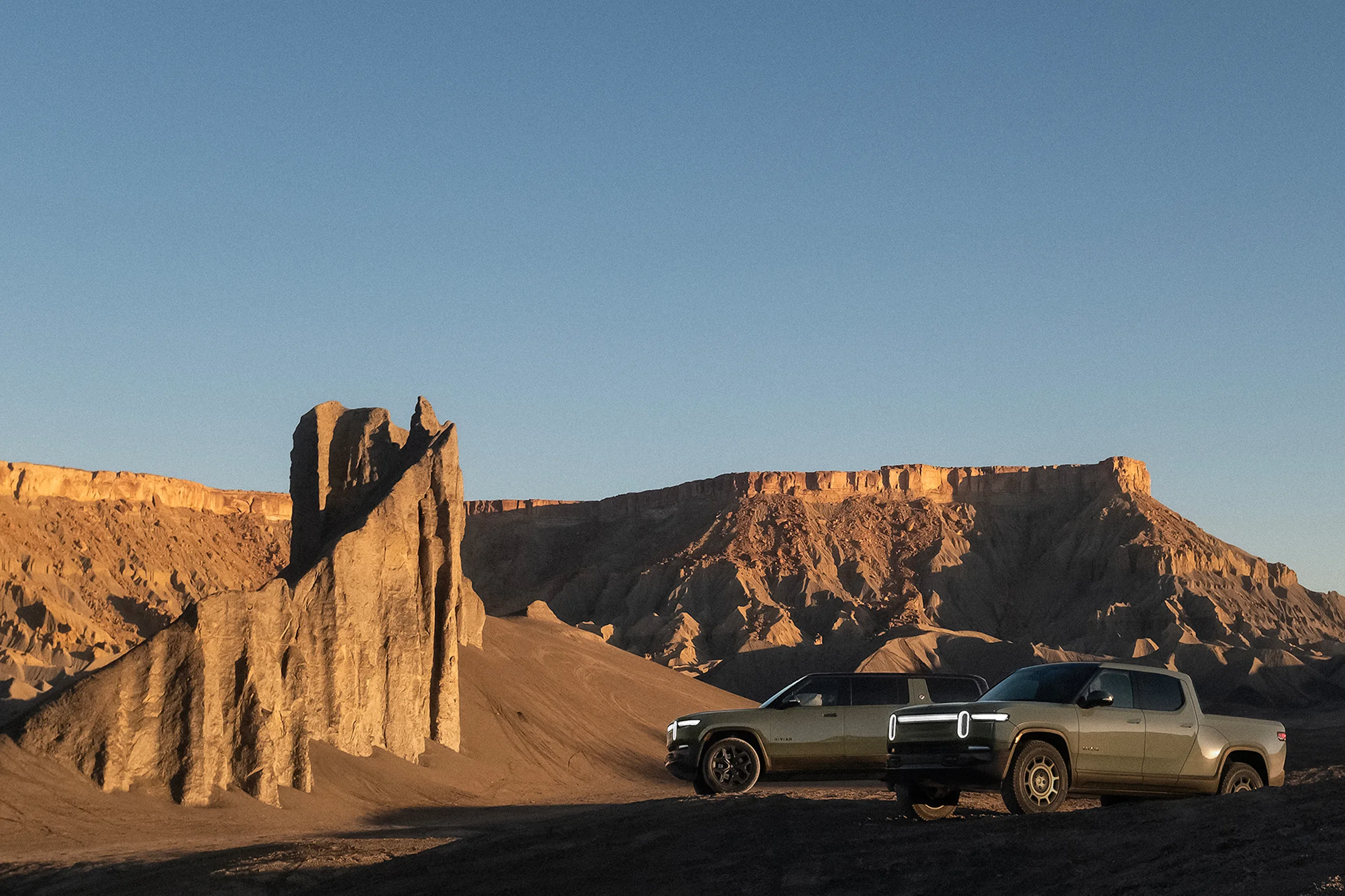Two Rivian electric vehicles parked in a canyon with red-rock cliffs extensively filling the background. One car is a Rivian R1S electric SUV an the other is Rivian R1T electric SUV. Both are in the limited edition Launch Green color.
