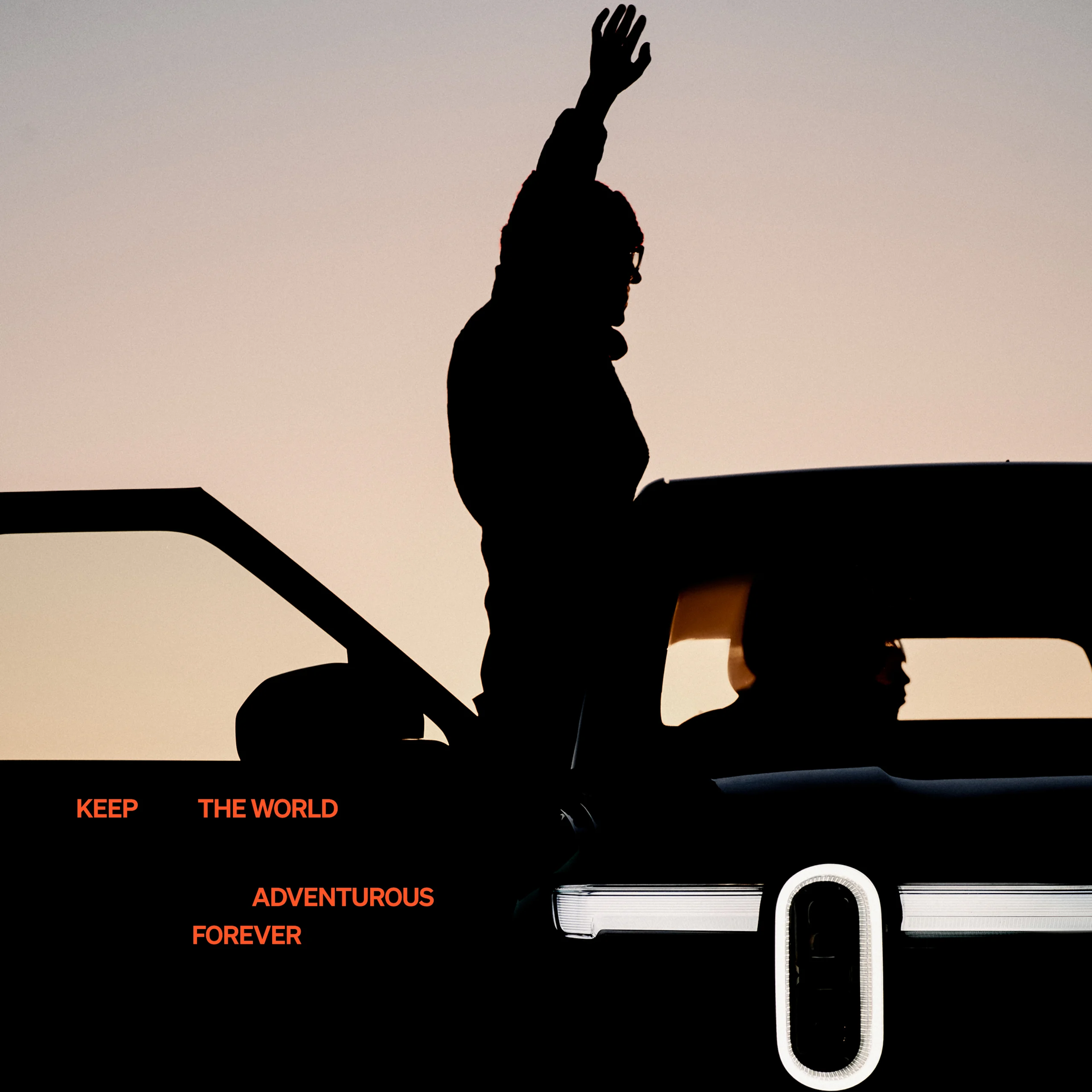 The silhouette of the front a Rivian electric vehicle as it is heavily shadowed by the setting sun. A the passenger door is open and person is standing, looking out towards the background.