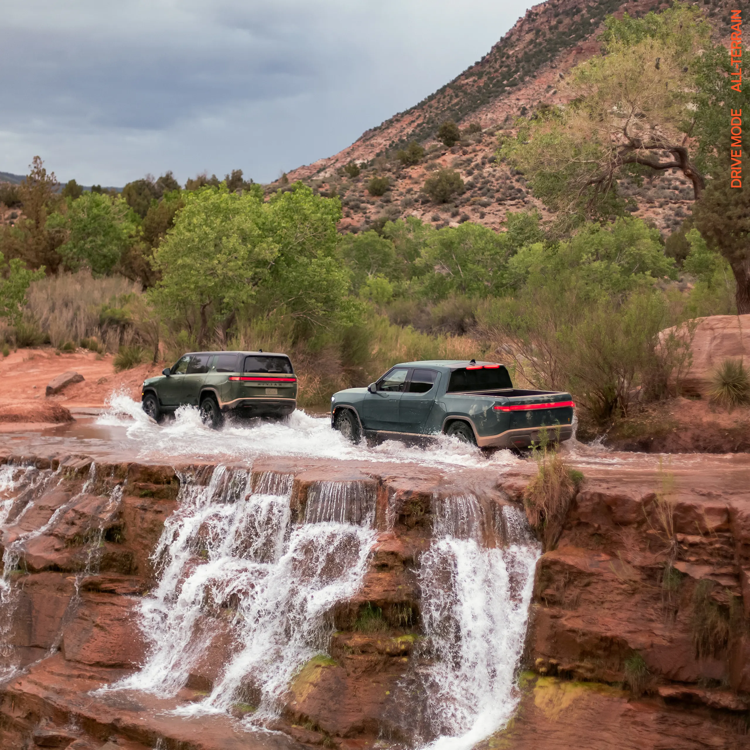Two Rivian electric vehicles driving through a heavy stream of water atop a red-rock cliff. One car is a Rivian R1S electric SUV with Forest Green paint, and the other is Rivian R1T electric SUV with Storm Blue paint.