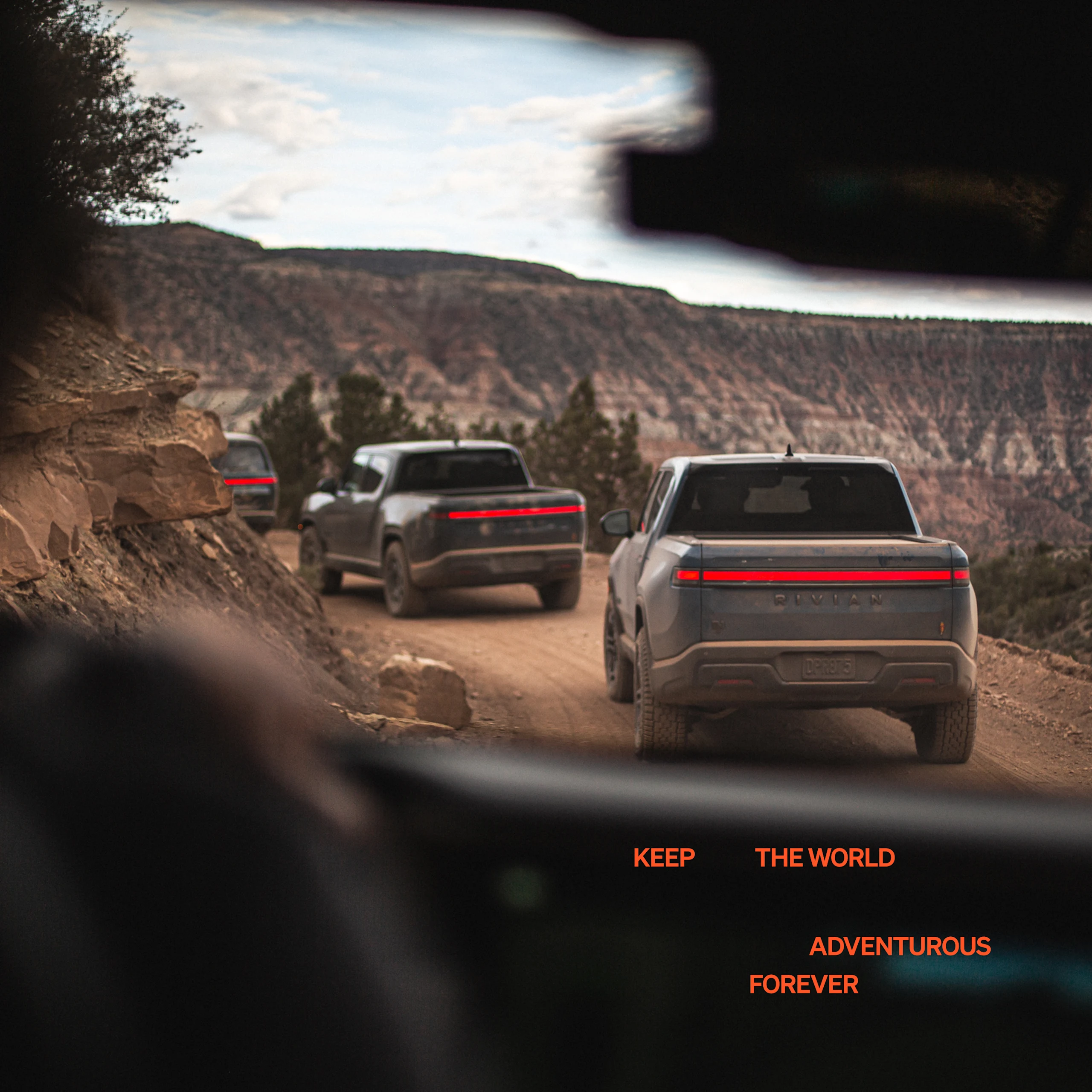 A line of three Rivian R1T electric trucks driving around a winding dirt road. The picture is taken from the inside of another Rivian vehicle that is trailing these three vehicles. 