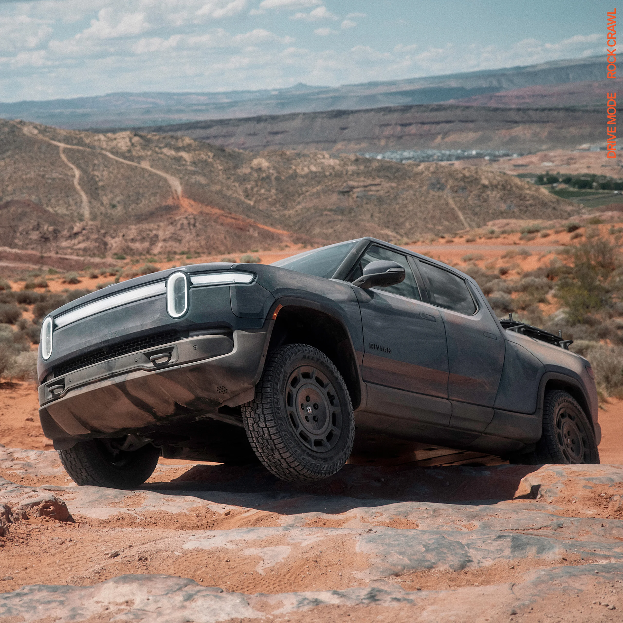A Rivian R1T electric truck crawling over rugged terrain. The front driver wheel is off the ground. The car features 20” Dune Satin Graphite All-Terrain wheels. 