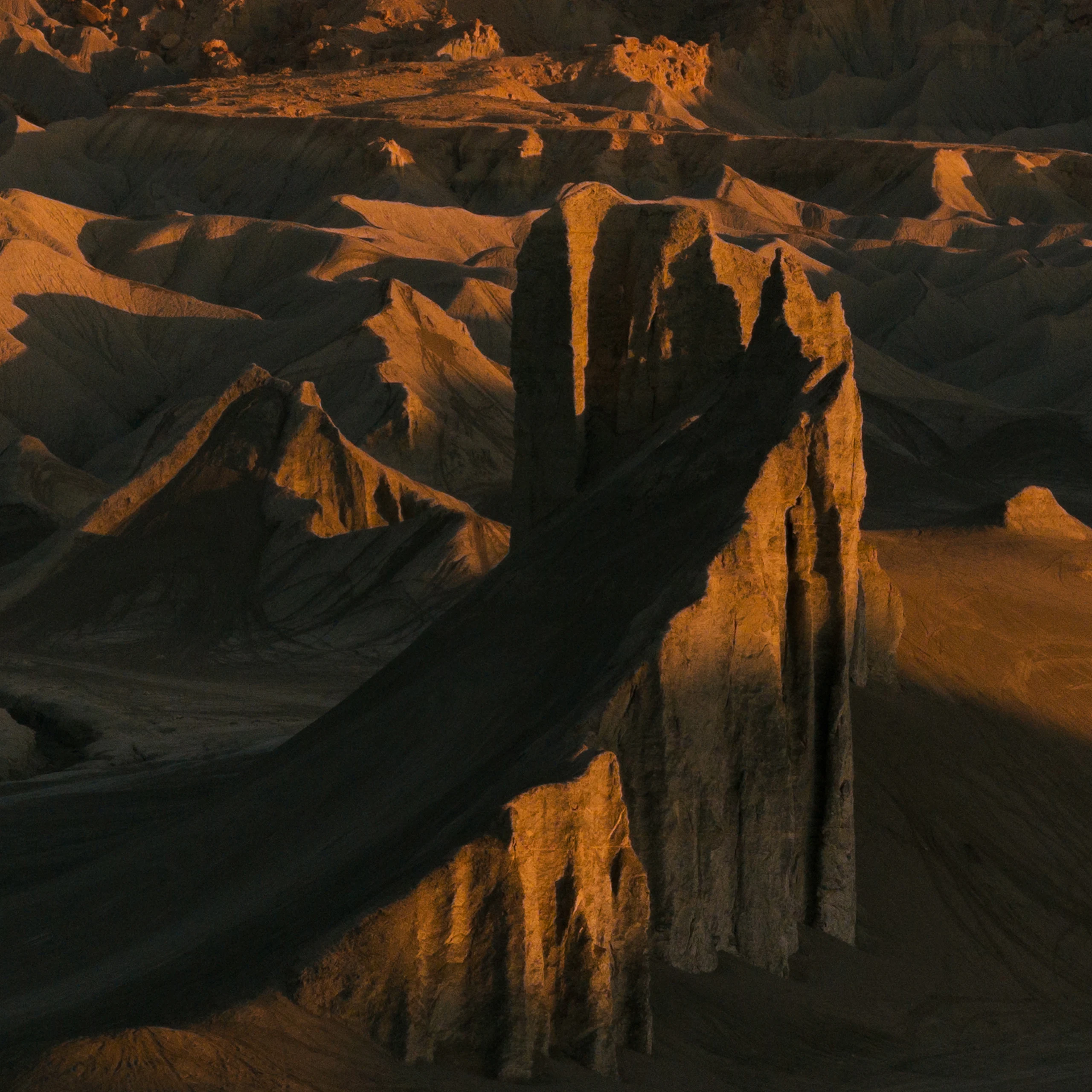 A massive slanted red rock cliff, with numerous other smaller hills in the background. The cliffs cast shadows in the sundown light.