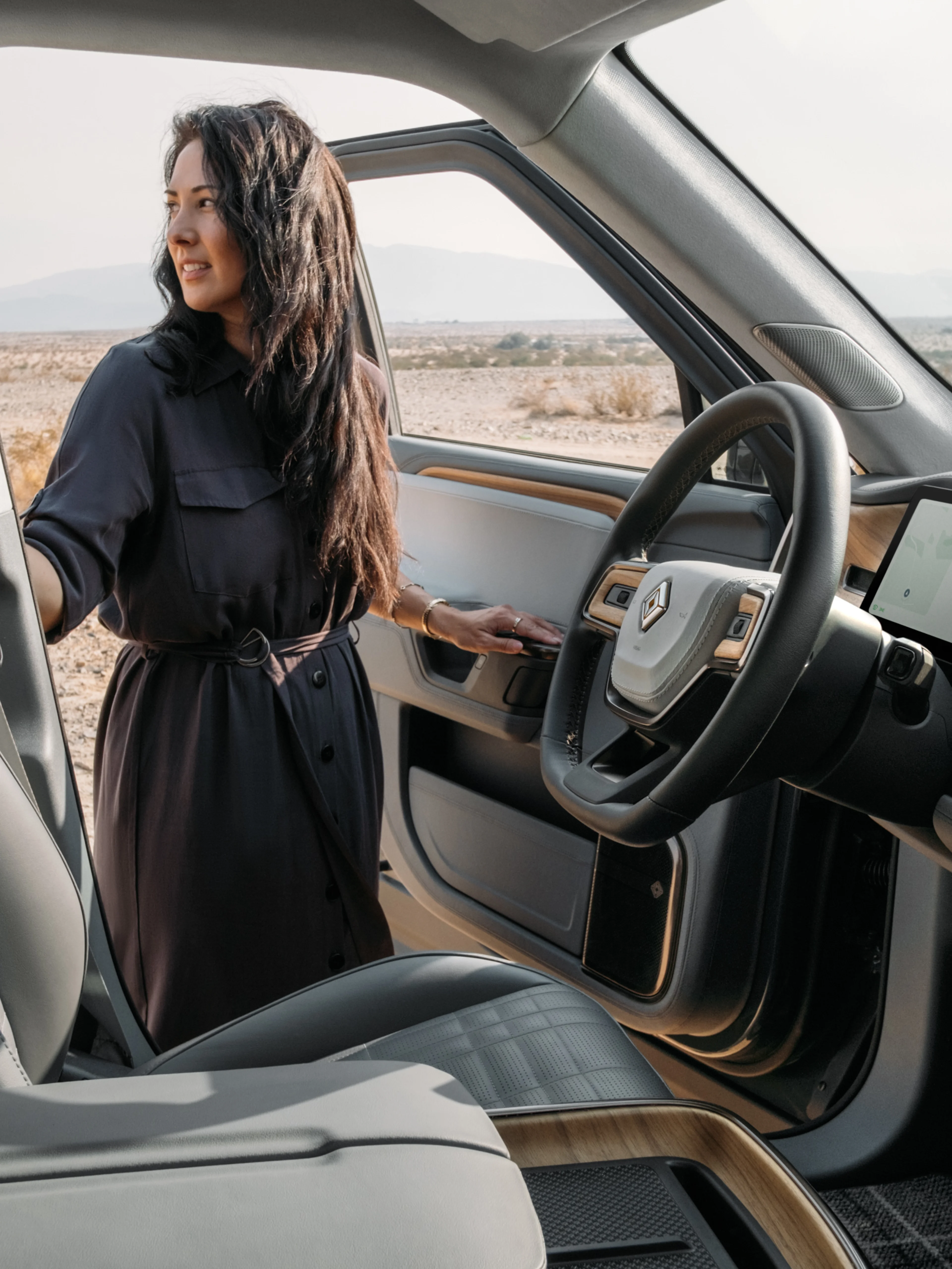 A woman in a dark dress standing beside the open driver door of a Rivian EV car.