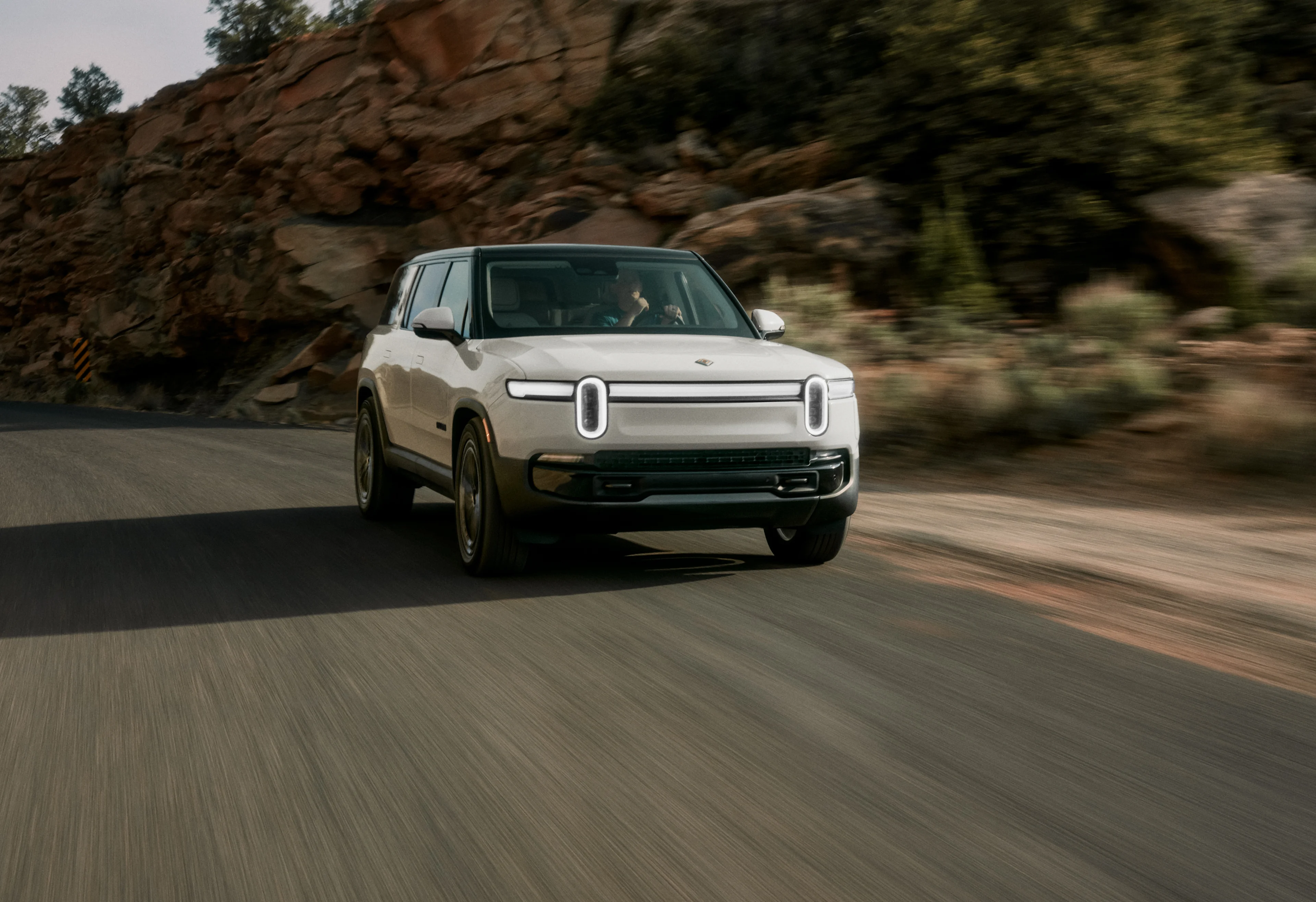 The front of a white Rivian Quad motor electric vehicle, swiftly driving down a open road with red-rock cliffs in the background.