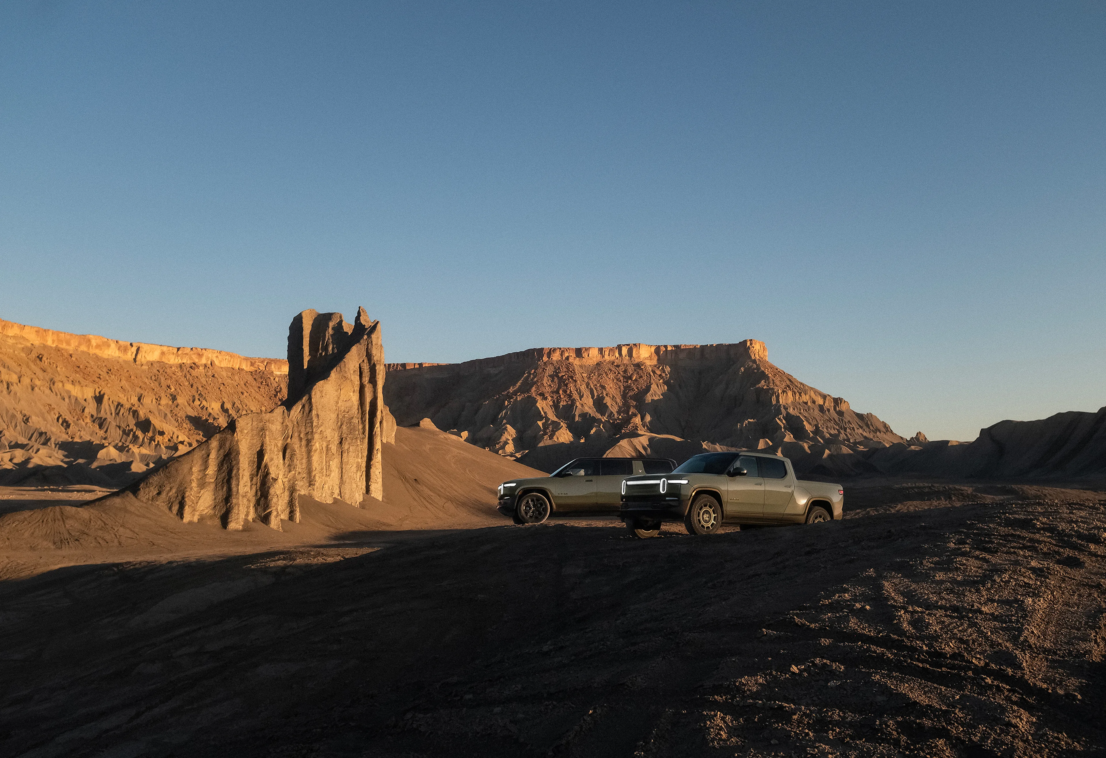Two Rivian electric vehicles parked in a canyon with red-rock cliffs extensively filling the background. One car is a Rivian R1S electric SUV an the other is Rivian R1T electric SUV. Both are in the limited edition Launch Green color.