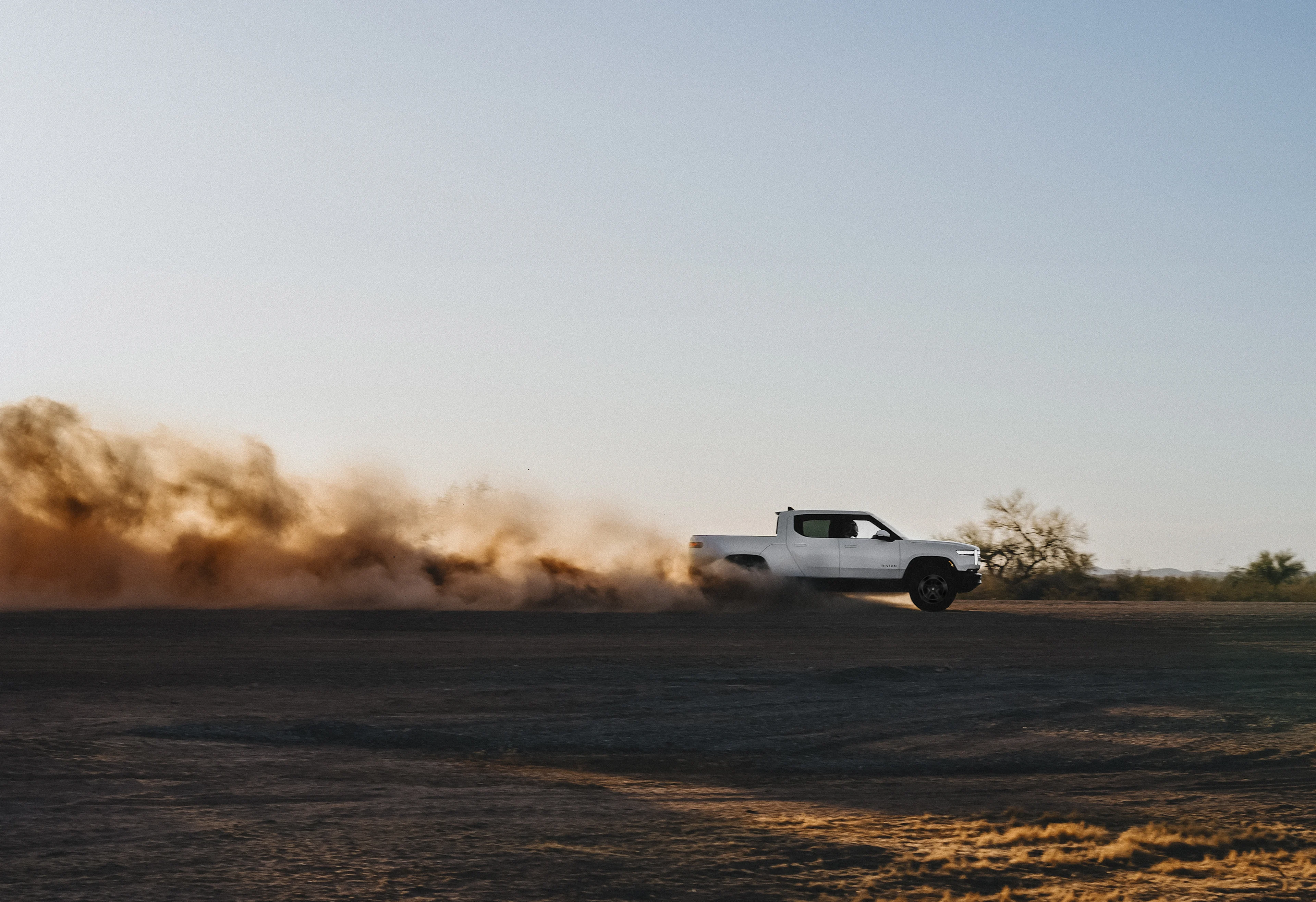 A white Rivian R1T electric truck driving swiftly through a dirt valley as the sun sets. A plume of dust kicks out of the back.