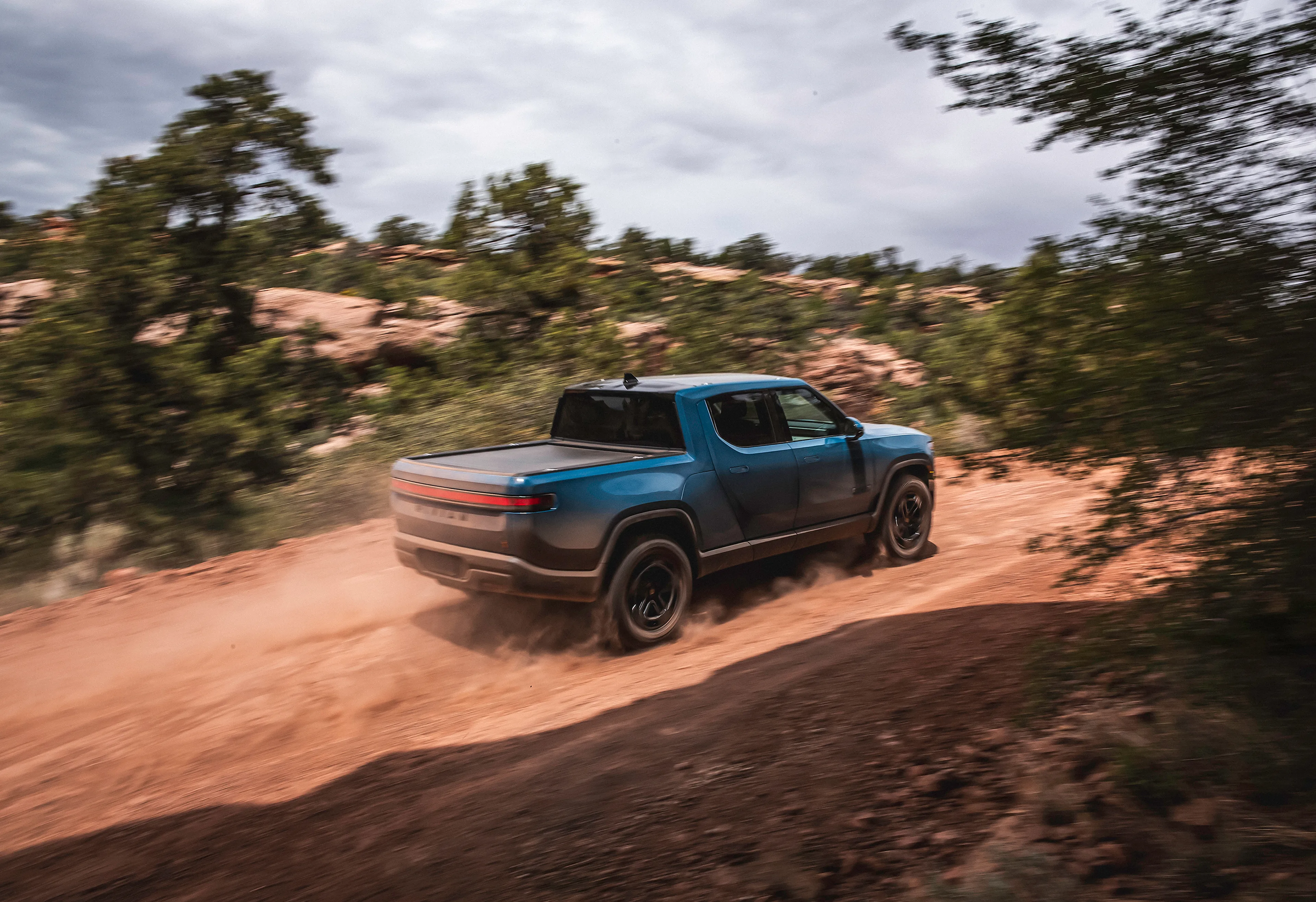 A backside view of a blue Rivian R1T electric truck driving up a dusty dirt road.