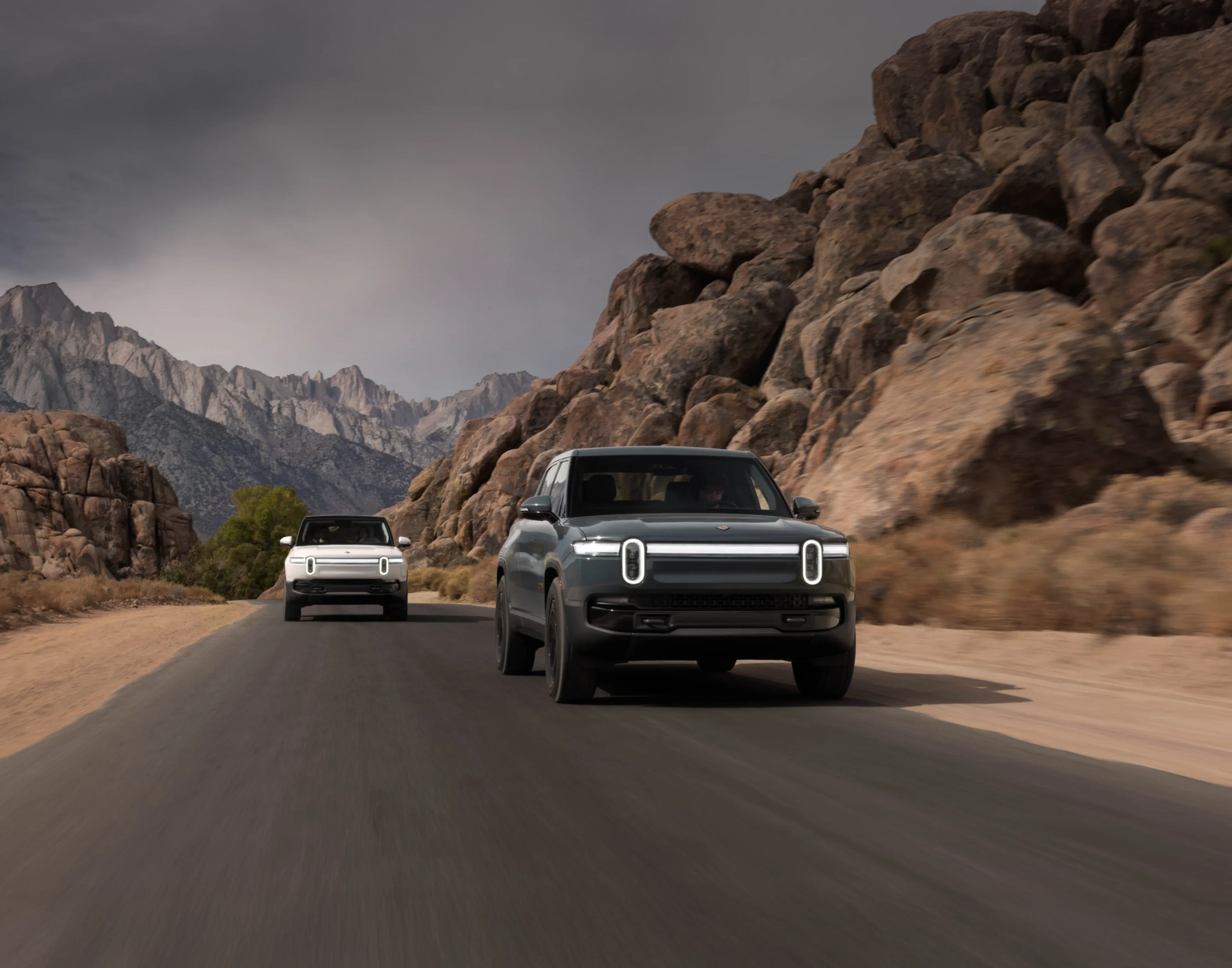 Two Rivian R1S electric SUVs driving through a scenic mountain road surrounded by rocky cliffs and cloudy skies.