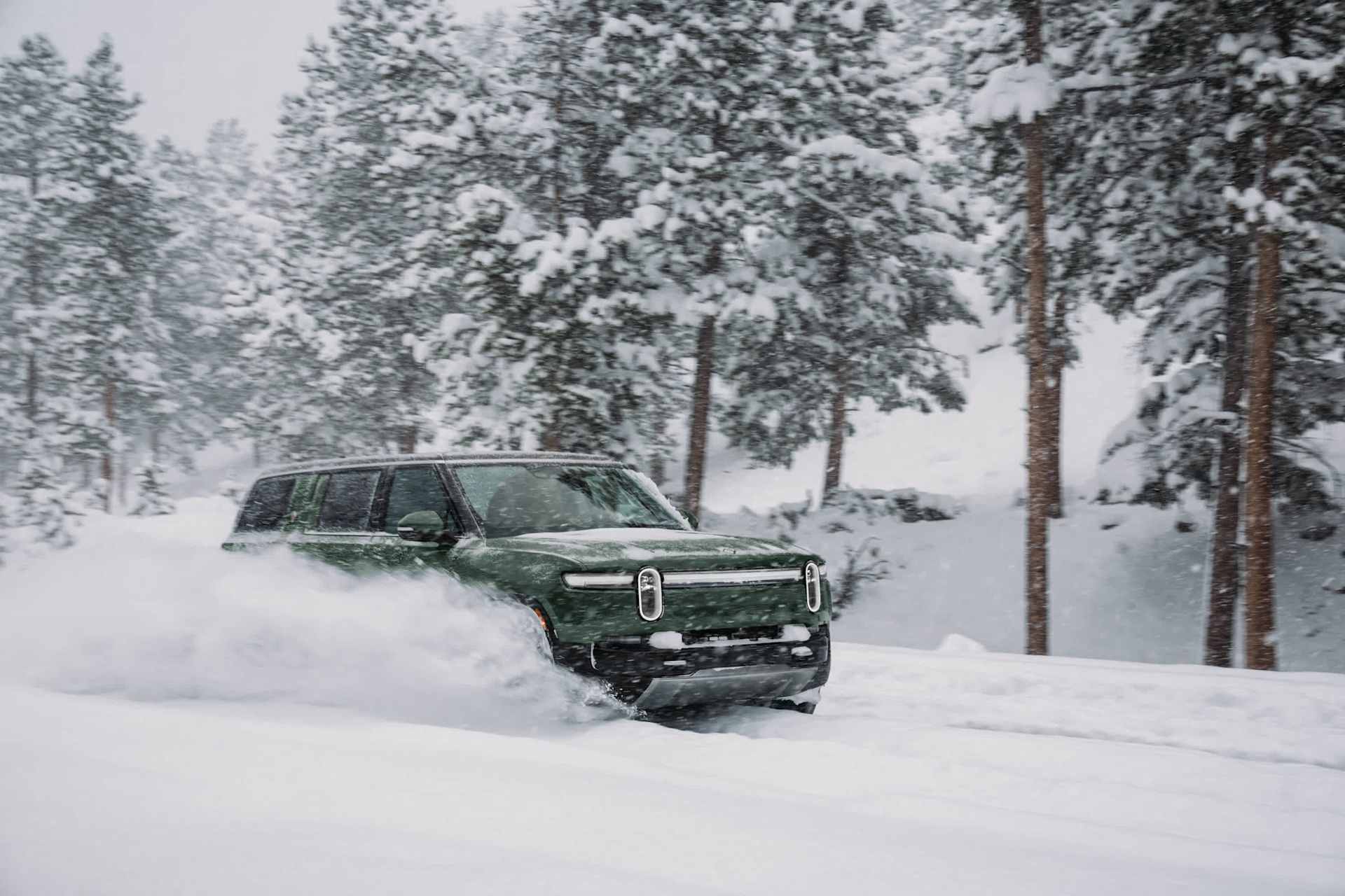 Green Rivian SUV driving through deep snow, kicking up a spray. Snow-covered pine trees are visible in the background.